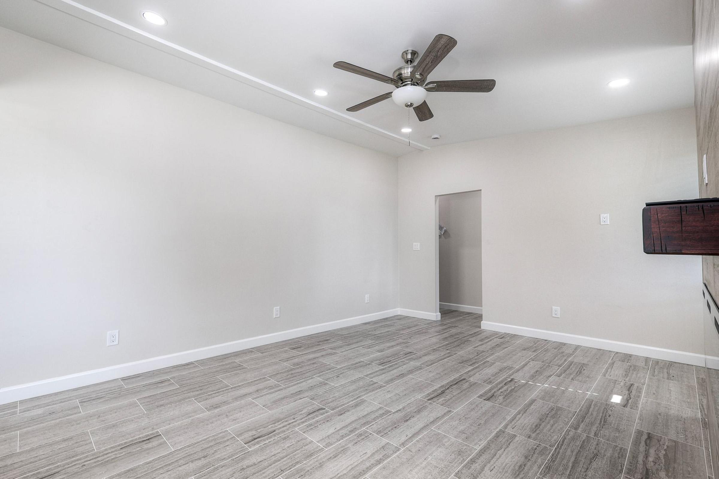 A spacious, modern living room featuring beige walls, a ceiling fan, and large gray tile flooring. The area is well-lit with several recessed ceiling lights and includes an open doorway leading to another room. The minimalist design creates a bright and inviting atmosphere.