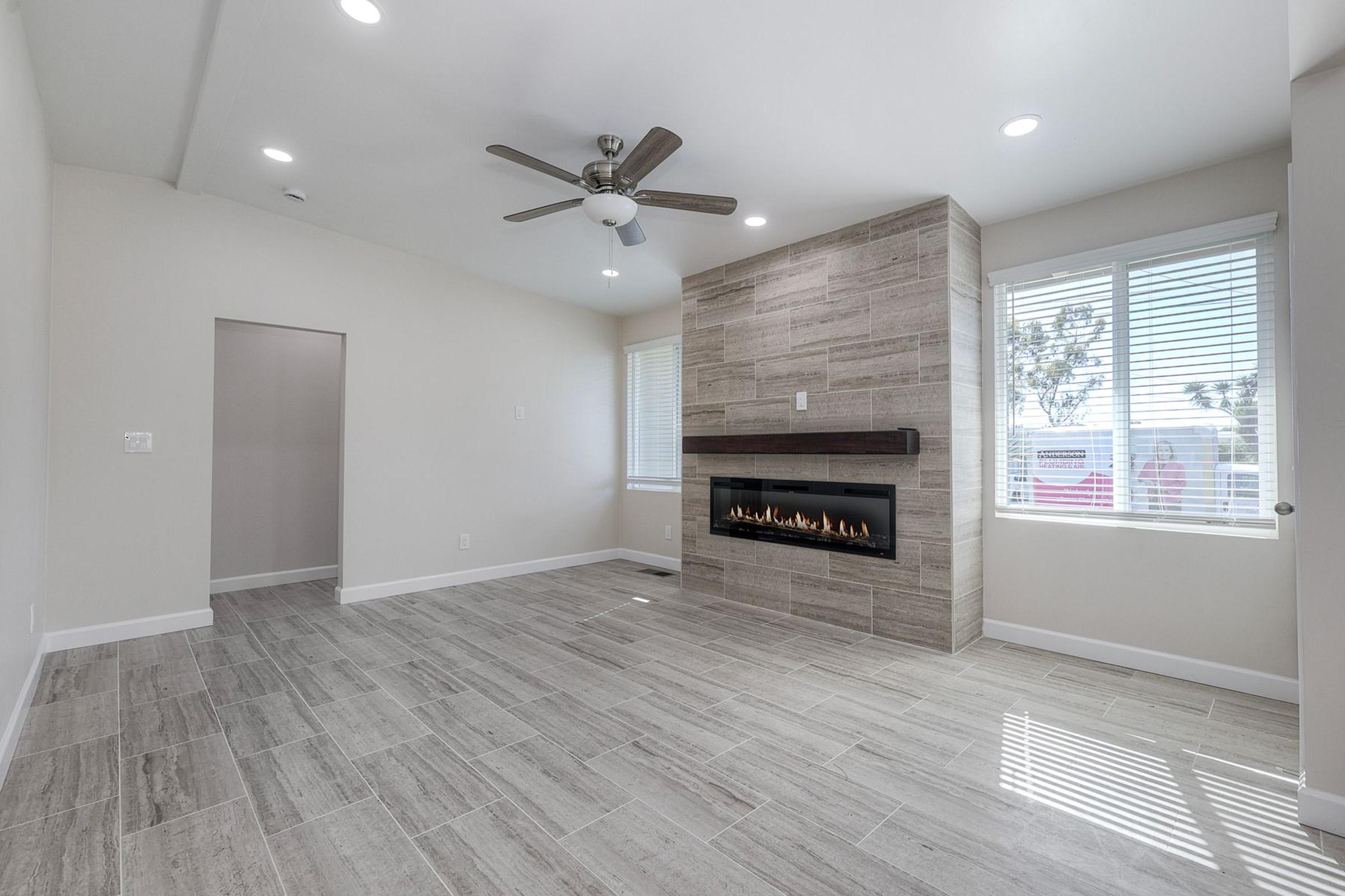 A modern living room featuring light-colored walls, a ceiling fan, and a tiled floor. The focal point is a stylish linear fireplace built into a stone wall. Large windows allow natural light to fill the space, creating an open and airy atmosphere. An adjacent room is partially visible.