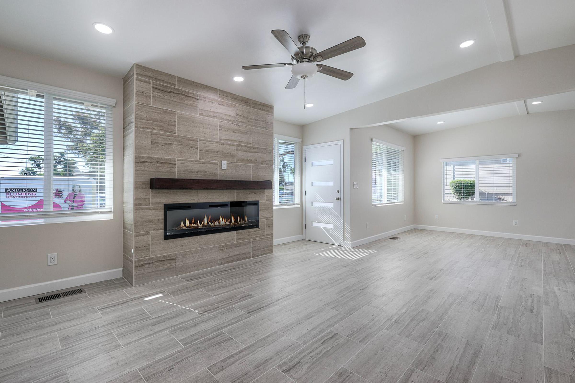 Bright and spacious living room featuring a modern electric fireplace with a sleek stone facade, large windows allowing natural light, and a ceiling fan for ventilation. The room has neutral-colored walls and stylish flooring, creating an inviting atmosphere.
