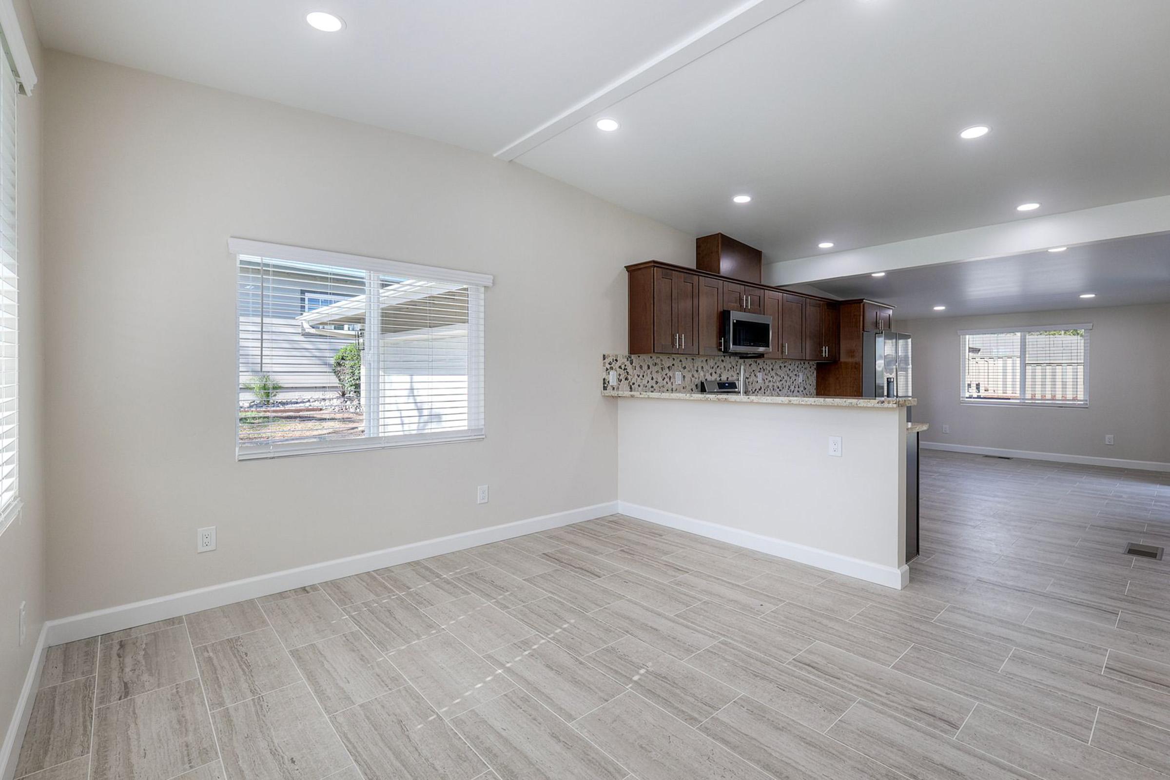 A spacious, modern interior view of a home featuring a light-colored living area with tiled flooring, large windows providing natural light, and a kitchen area with dark wooden cabinetry and a mosaic backsplash. The layout is open, enhancing the sense of space.
