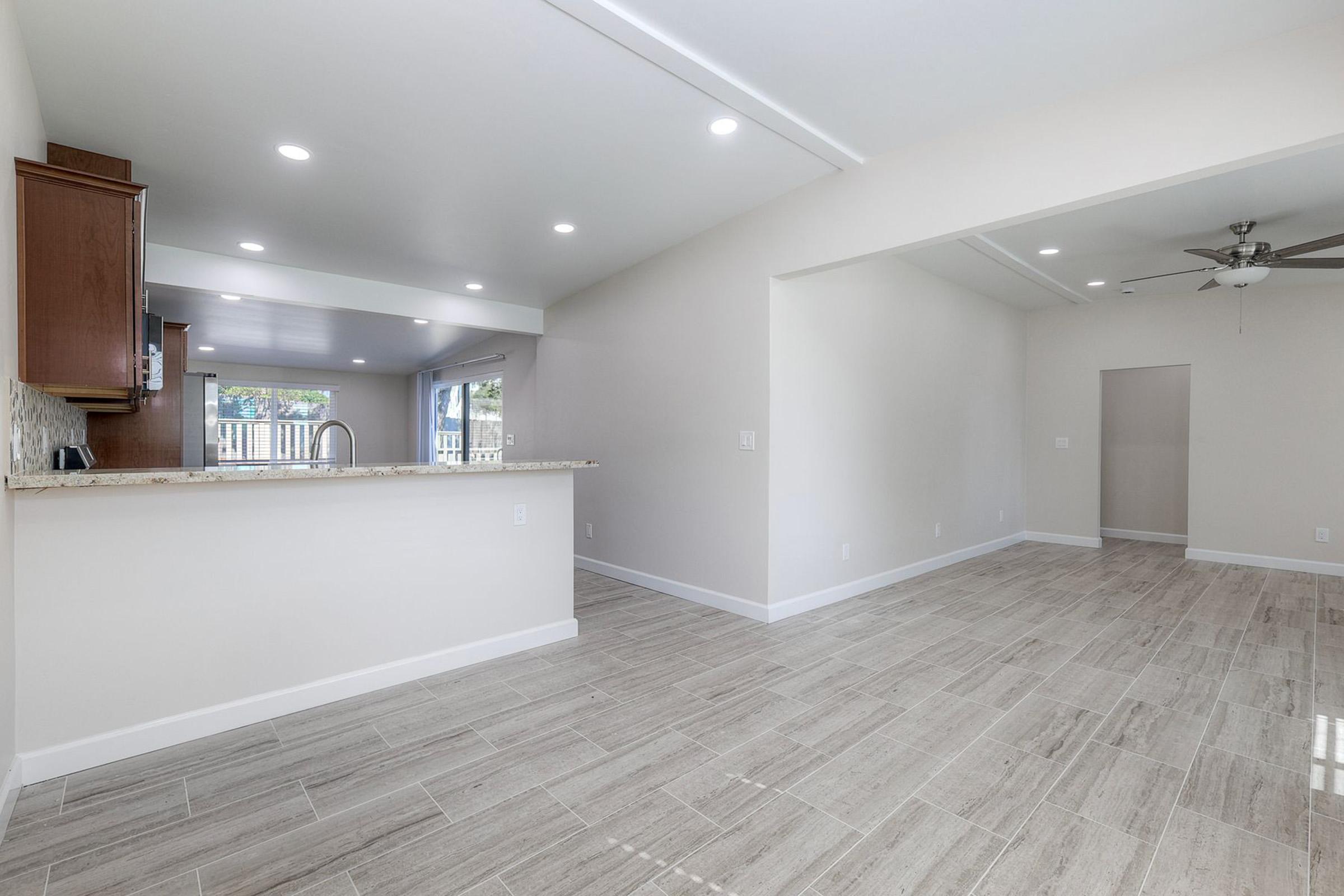 Modern open-concept living space featuring a kitchen with wooden cabinets and a breakfast bar. The light-filled area has tiled flooring, neutral-colored walls, and a ceiling fan. Large windows offer a view to the outdoors, enhancing the inviting atmosphere.