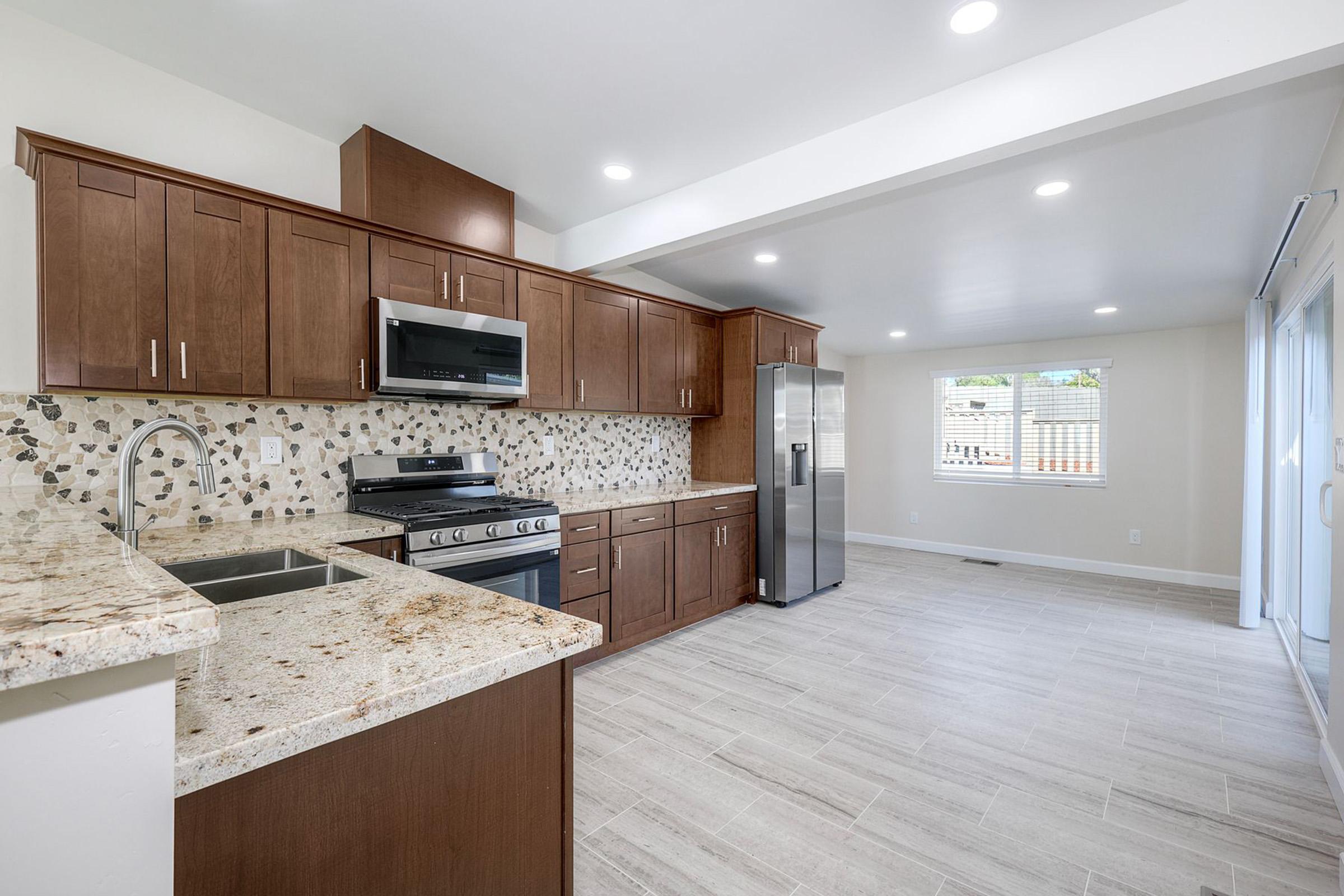 Modern kitchen featuring dark wood cabinetry, stainless steel appliances, and a granite countertop. The backsplash consists of decorative tiles, and large windows allow natural light to fill the space. A sliding door leads to an outdoor area, while the floor is covered in light, tile-style flooring.