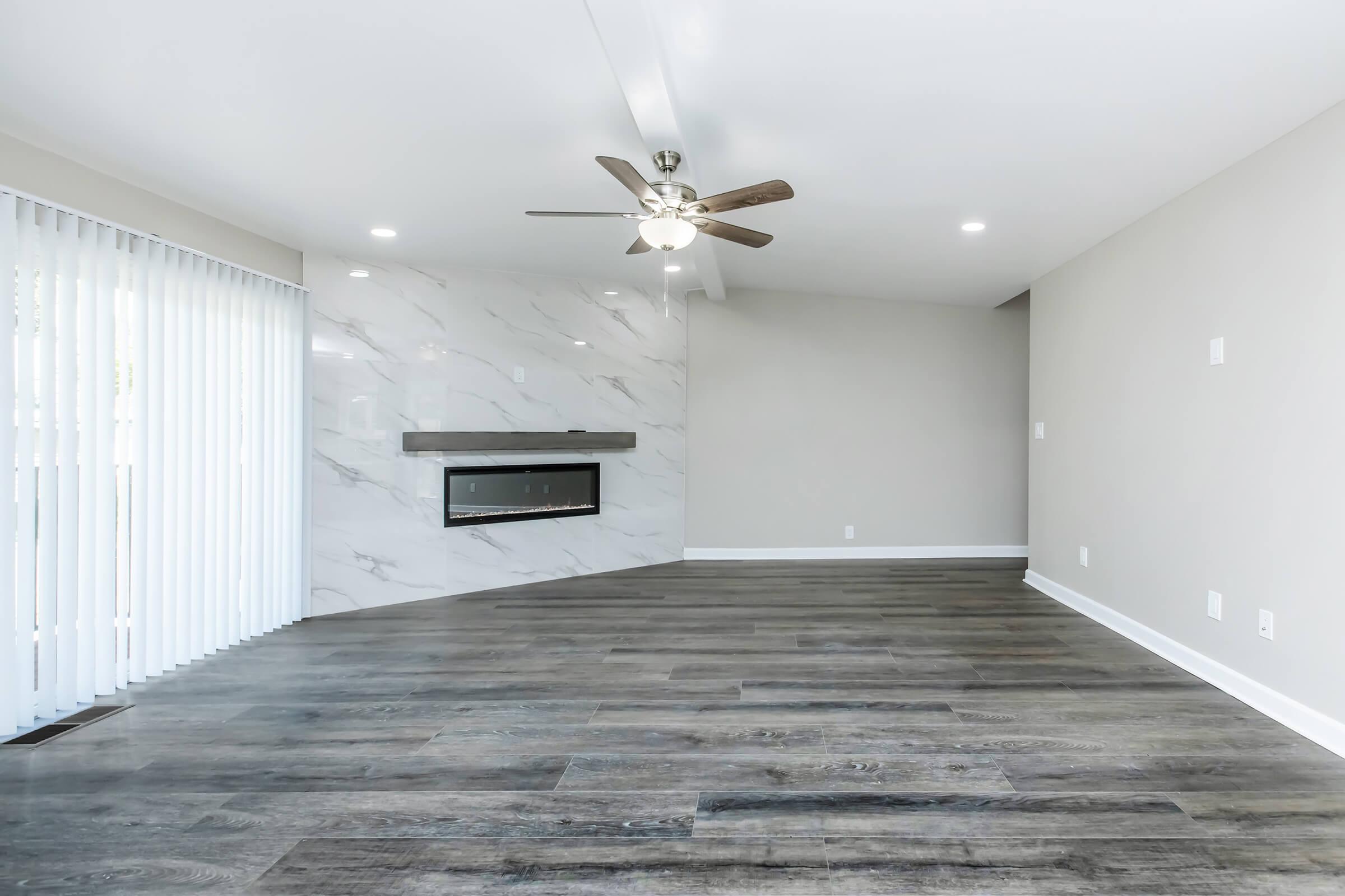 Spacious living room featuring a modern design with light wood-style flooring, a ceiling fan, and a minimalist fireplace adorned with gray marble. Large sliding glass doors allow natural light, and there are soft white walls creating a bright, airy atmosphere.