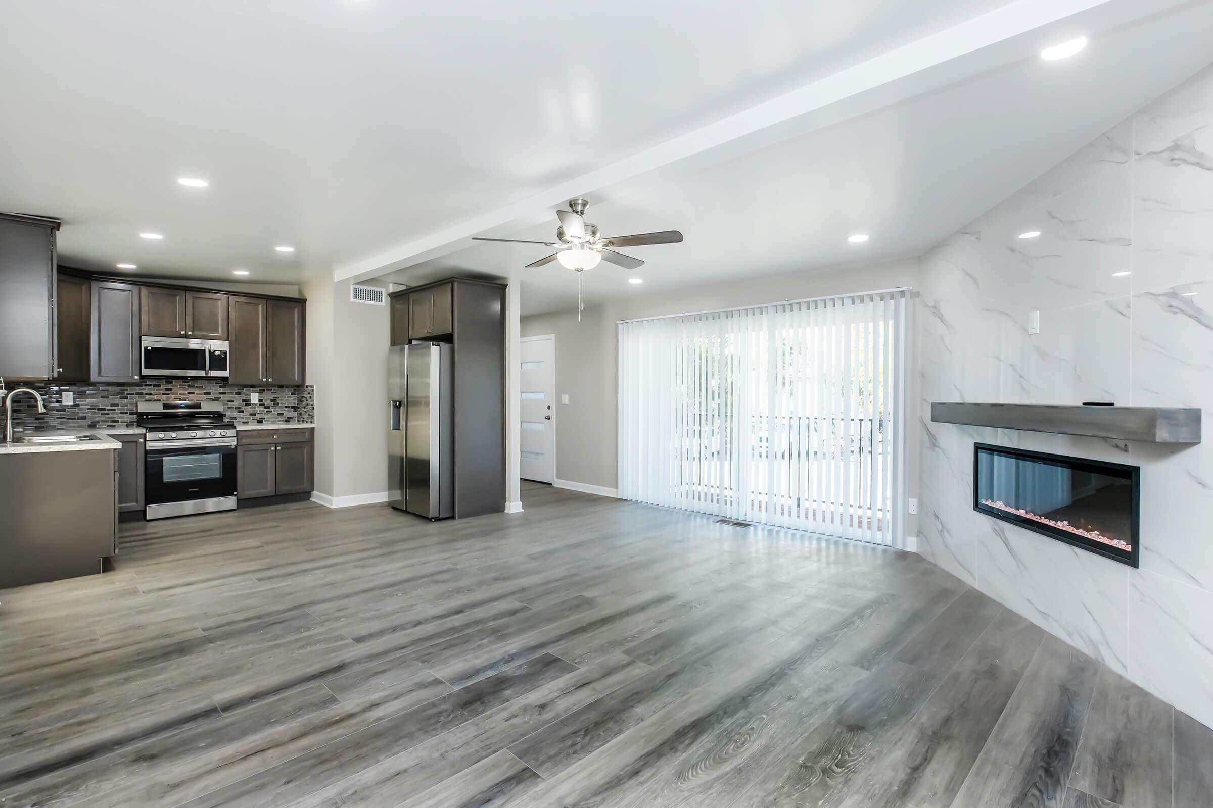 Modern open-concept living space featuring a stylish kitchen with stainless steel appliances, a ceiling fan, and sleek cabinetry. The room has large windows with vertical blinds and a contemporary electric fireplace, set against light marble accents on one wall, and hardwood flooring throughout.