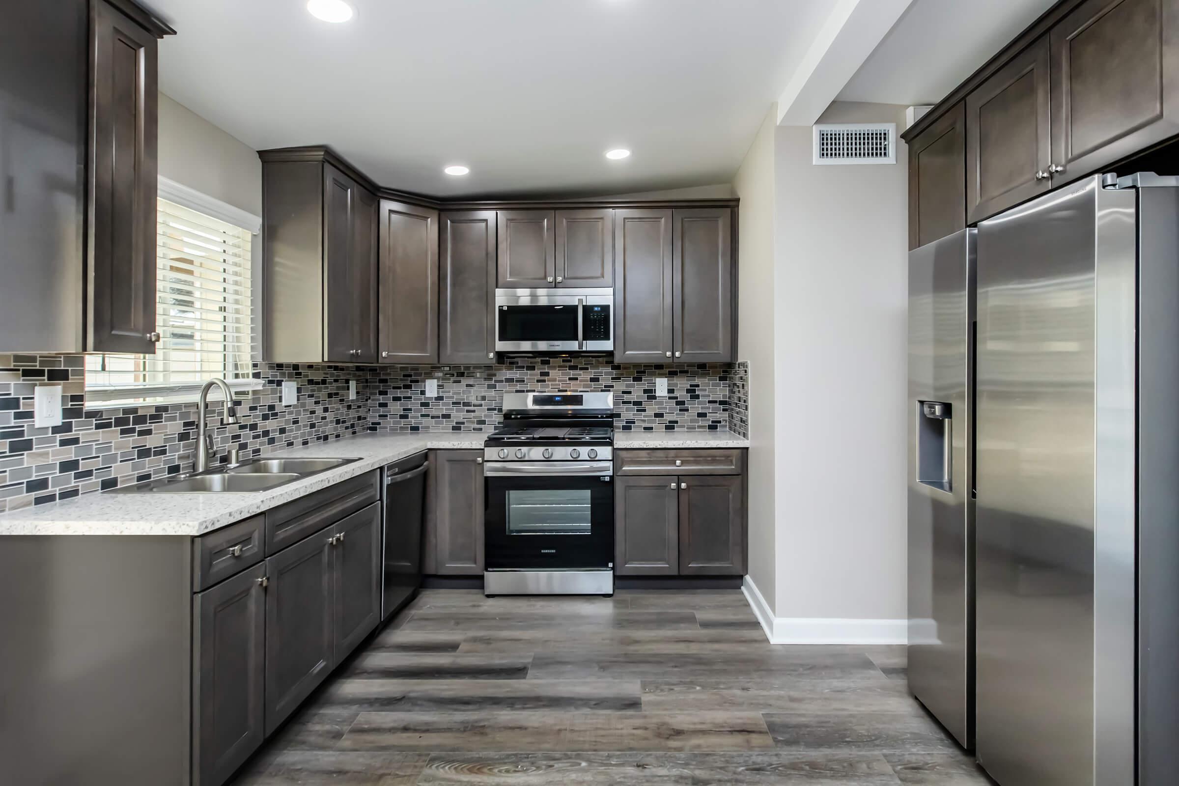 Modern kitchen featuring dark wood cabinets, a stainless steel refrigerator, and a stove with an oven. The countertop is made of light-colored stone, and the backsplash is composed of mosaic tiles in various shades. Natural light enters through a window, illuminating the space with a warm ambiance.