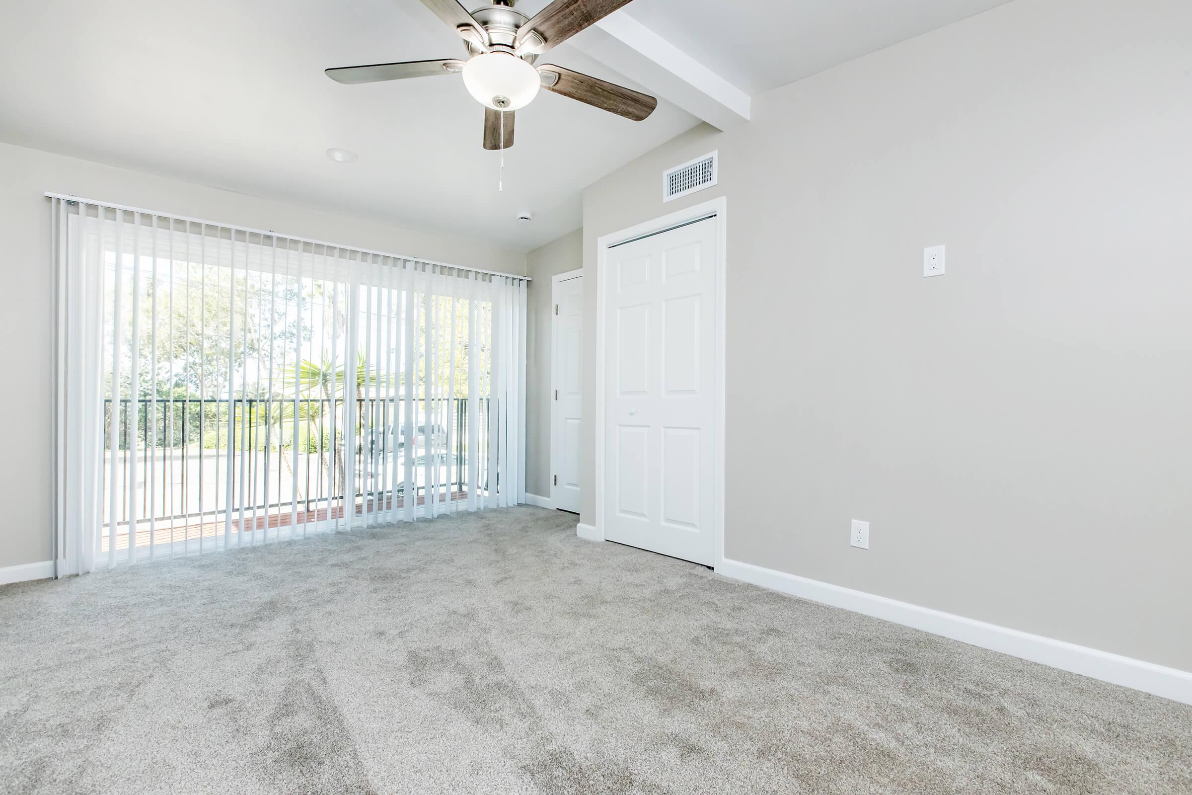 A spacious, modern room featuring light gray walls and plush carpeting. A ceiling fan provides light and air circulation. Glass sliding doors with vertical blinds lead to a small balcony area, enhancing natural light. A white closet door is visible on the right. The overall atmosphere is bright and airy.