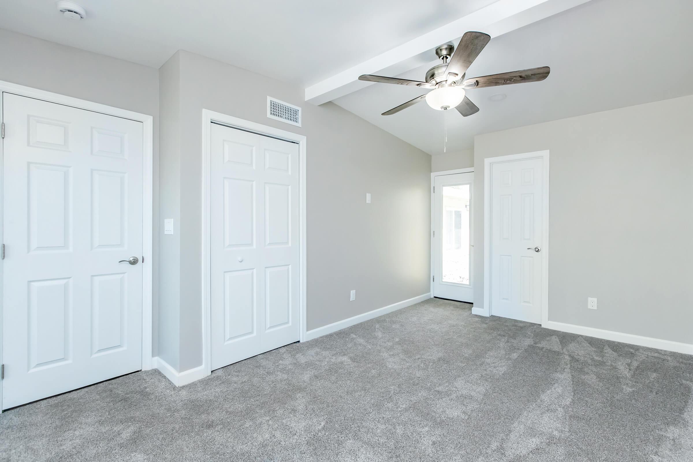 Spacious room with light gray walls and plush carpet, featuring two white closet doors and a ceiling fan. A door leads to another area, and natural light enters through a window. The room has a clean, modern aesthetic with minimal decor.