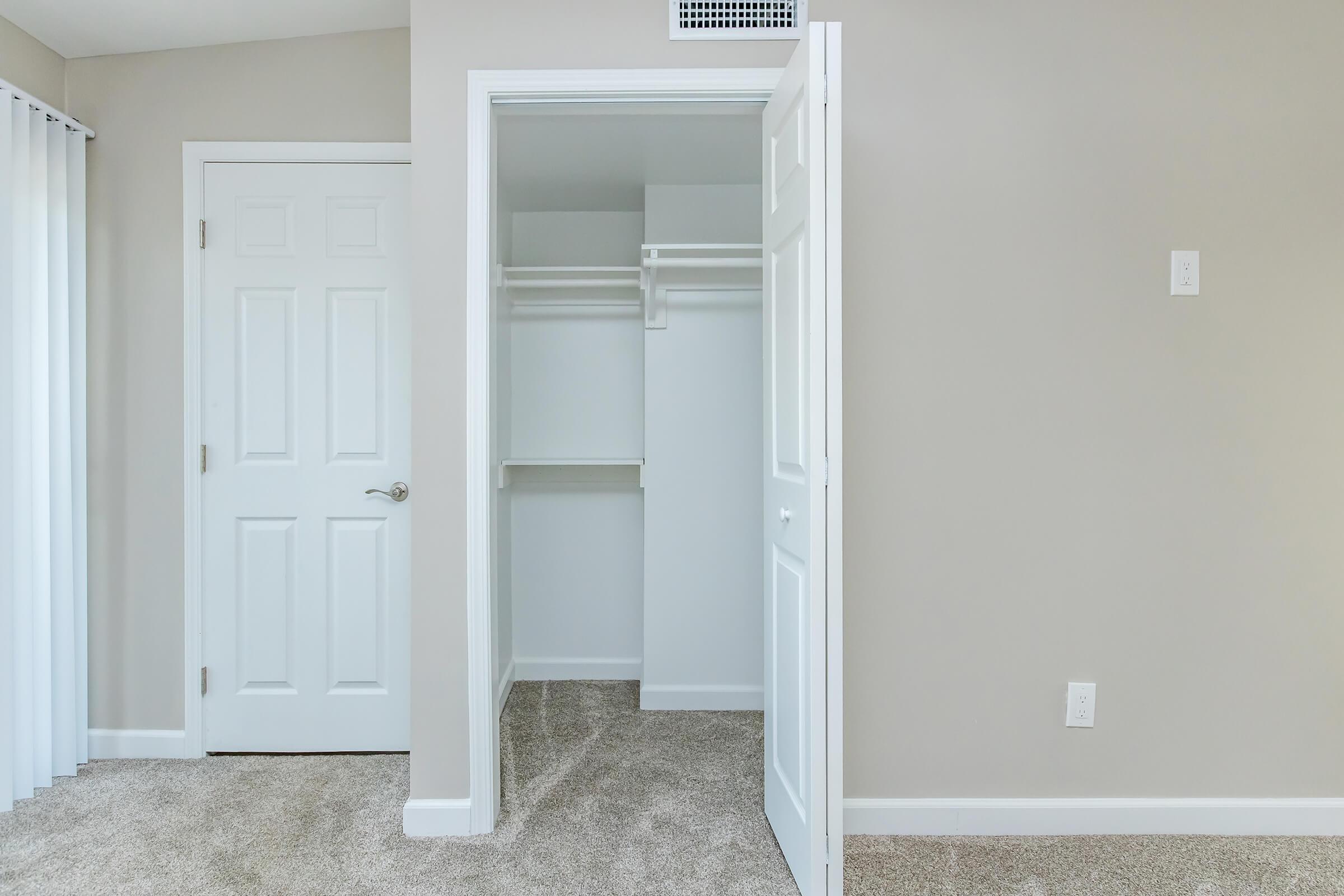 Empty room featuring a beige wall and carpeted floor. A closet with white shelves is visible, along with a closed door leading into the room. Natural light filters in from a nearby window, creating a bright and spacious atmosphere.