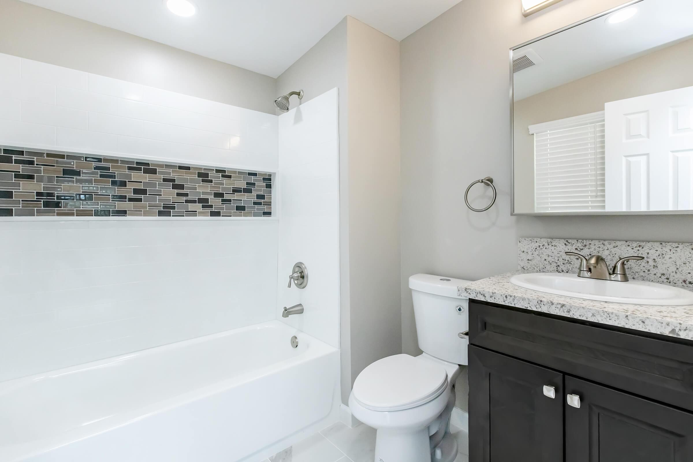 Modern bathroom featuring a white bathtub with a decorative mosaic tile strip, a sleek sink with a granite countertop, a toilet, and a large mirror. The walls are painted in a light color, and natural light streams in through a window. Elegant fixtures and clean lines create a contemporary aesthetic.