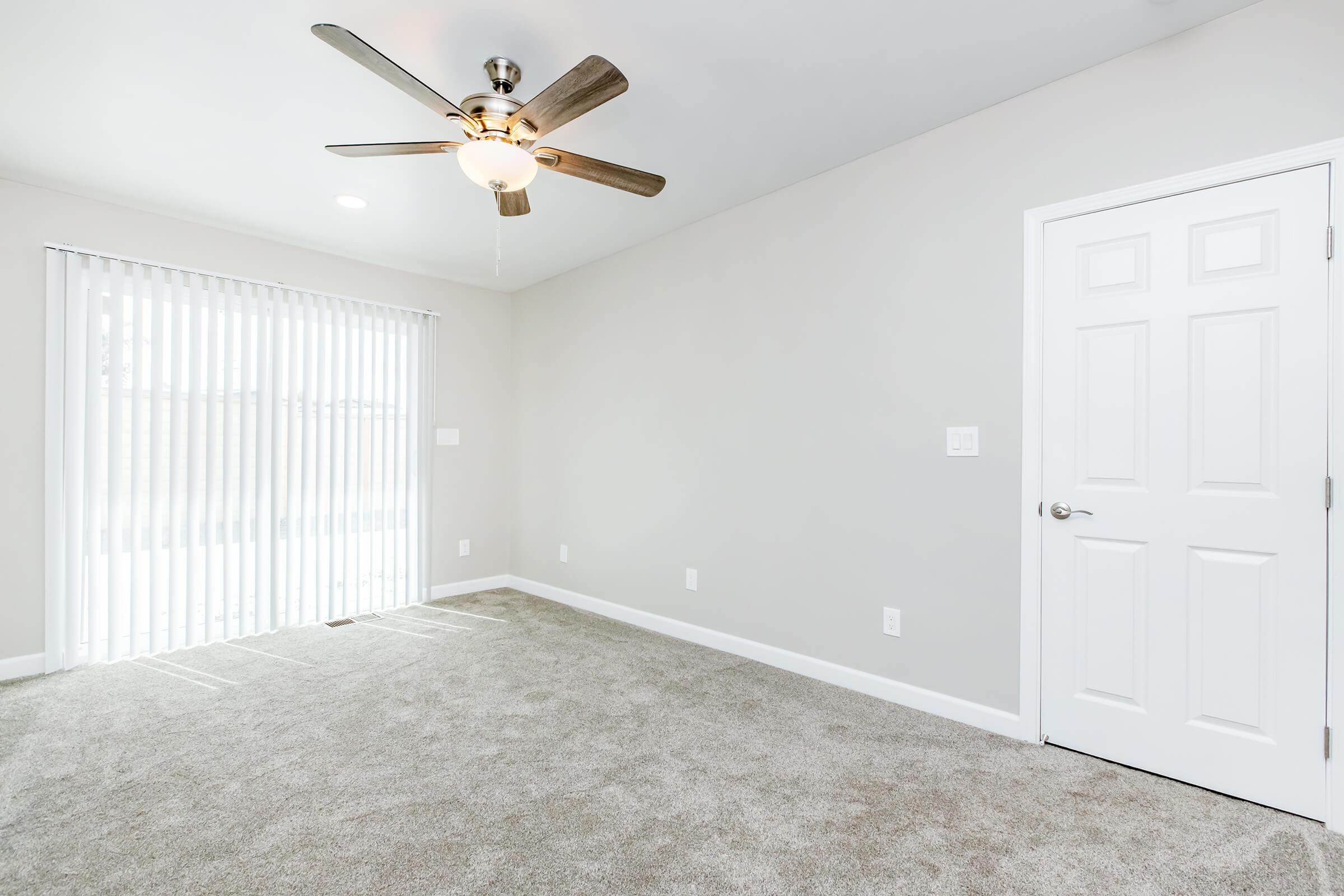 Empty room with light gray walls and carpeted floor, featuring a ceiling fan. A door is visible on the right side, and vertical blinds cover a window in the background, allowing natural light to enter. The space is minimal and bright, suitable for various interior designs.