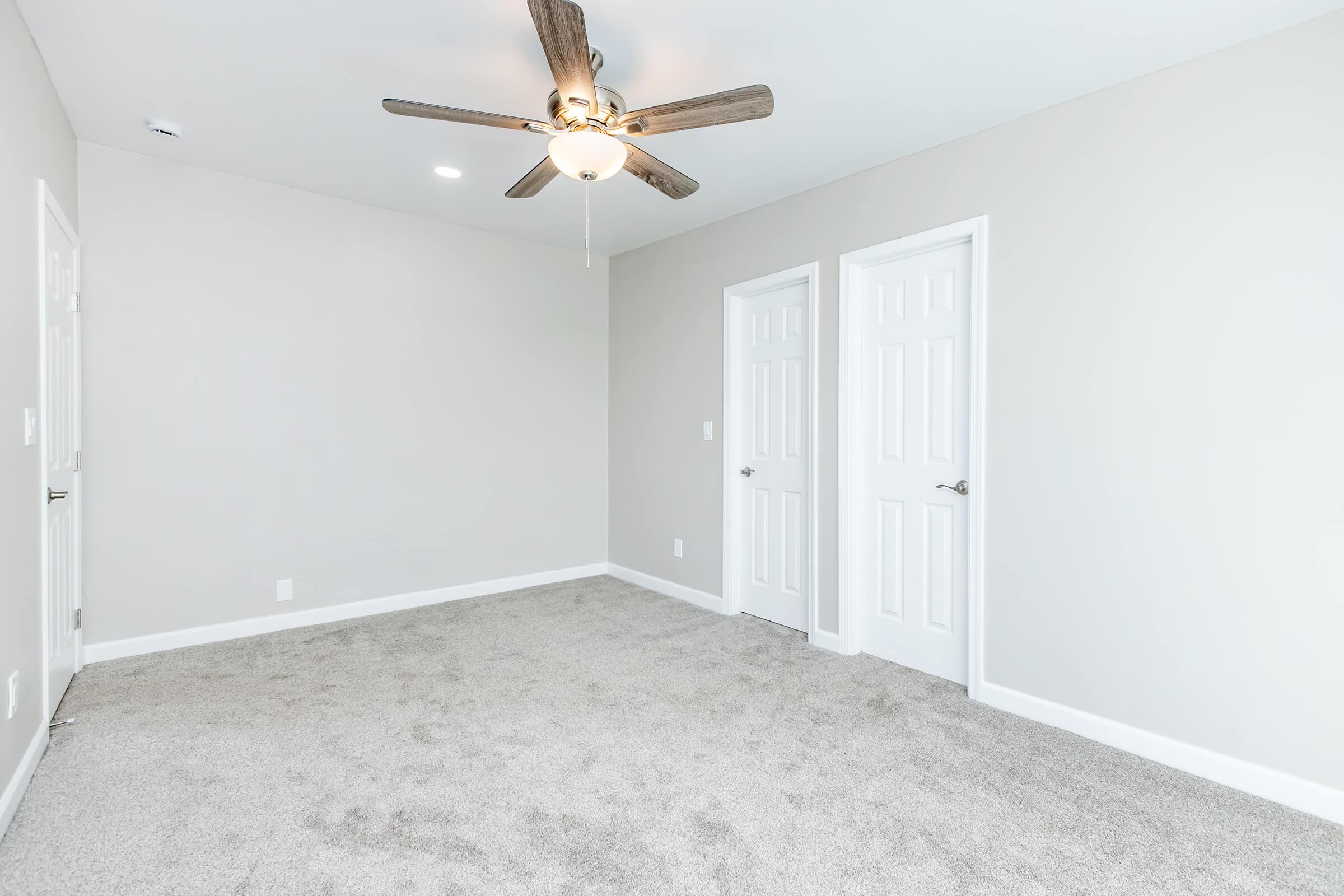 A spacious, empty room with light gray walls and a ceiling fan. There is plush carpet flooring in a neutral tone. Two white doors are visible on the right, and a single light fixture is on the ceiling, providing bright illumination. The overall ambiance is clean and modern.