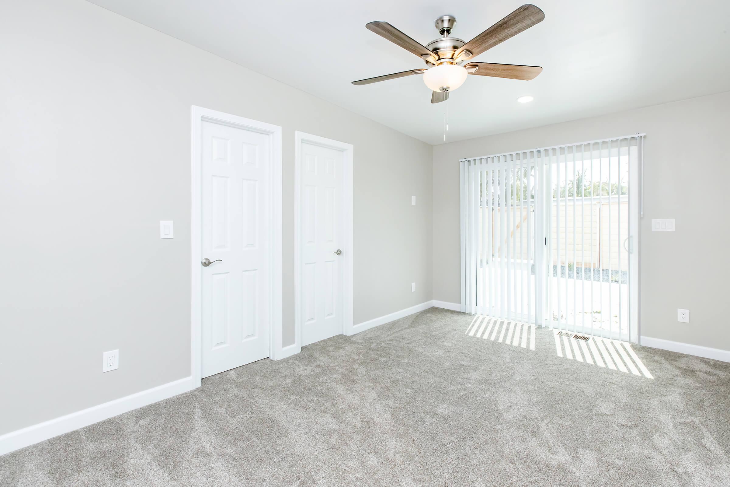 A modern, empty room with light grey walls and a ceiling fan. The floor is covered in plush, beige carpet. Two closed white doors are on the left, while large sliding glass doors on the right open to a patio, allowing natural light to enter. Vertical blinds hang over the glass doors.