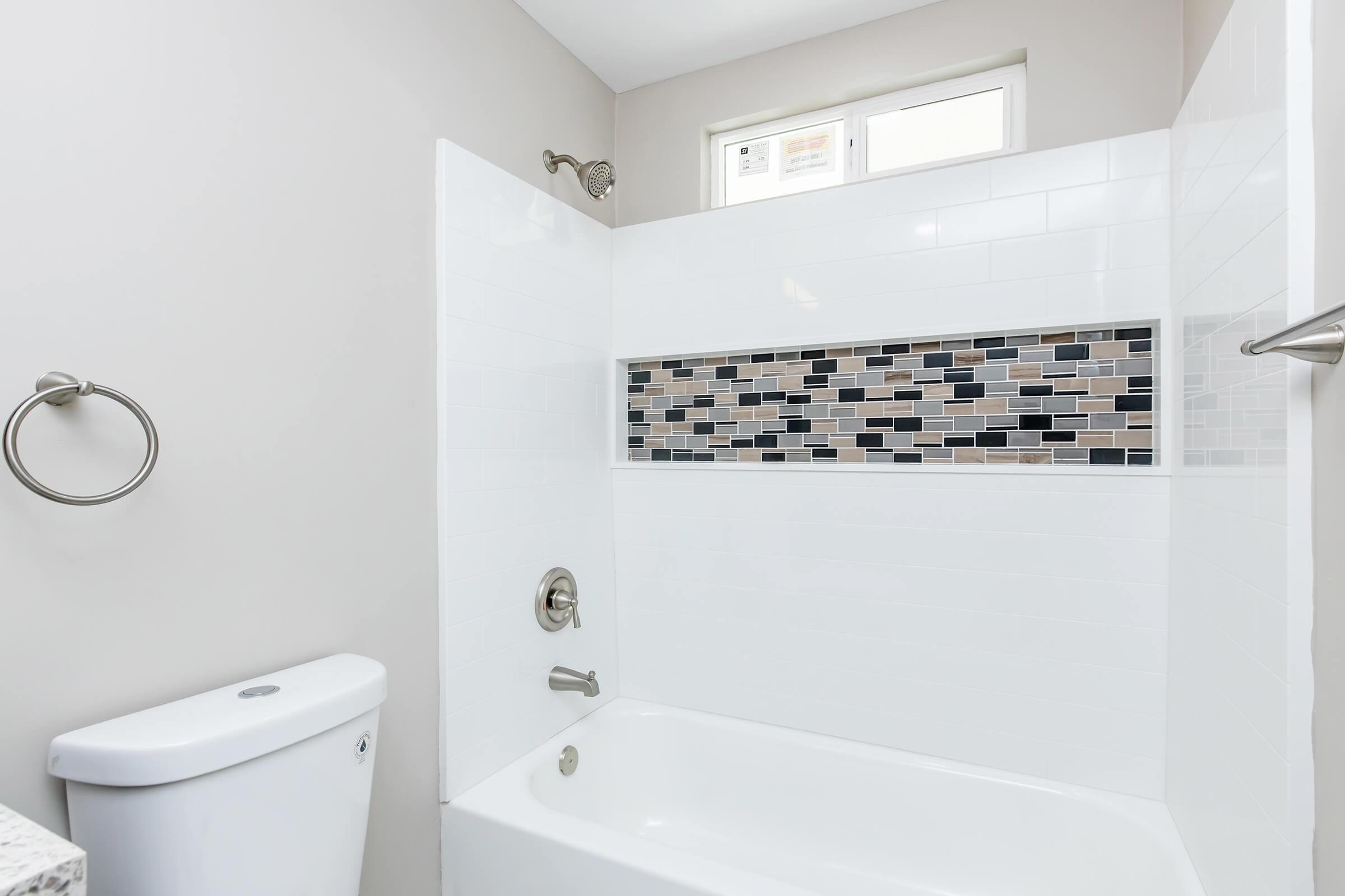 A clean and modern bathroom featuring a white bathtub with a showerhead above, light gray walls, and a decorative tile strip with black, gray, and white tiles. A toilet is positioned to the left, and there is a towel ring on the wall nearby. Natural light enters through a window.