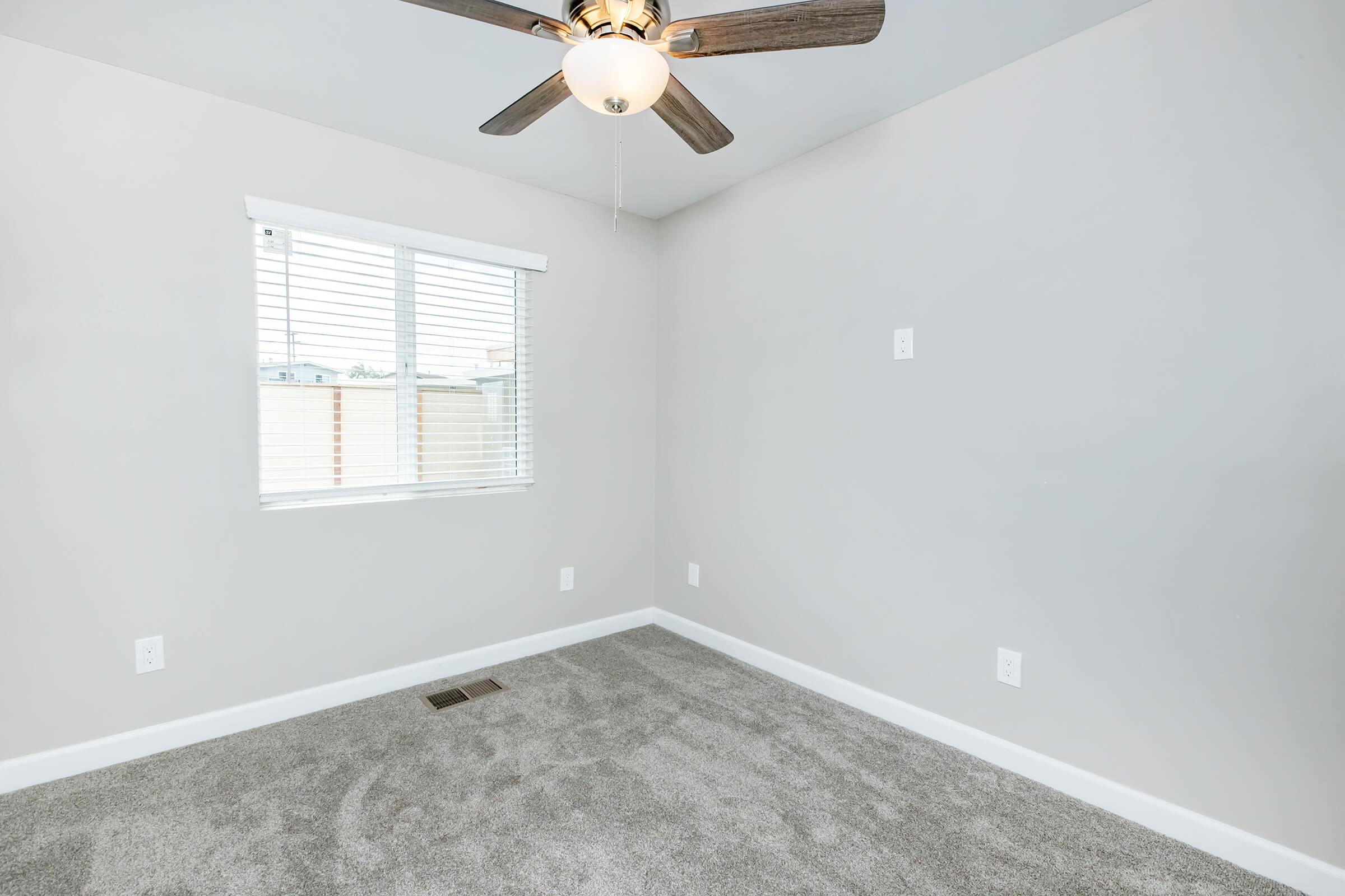 A light-filled room with gray walls and carpet. It features a ceiling fan with five blades, a window with white blinds, and electrical outlets on the walls. The space is empty, suggesting it could be used as a bedroom or office.
