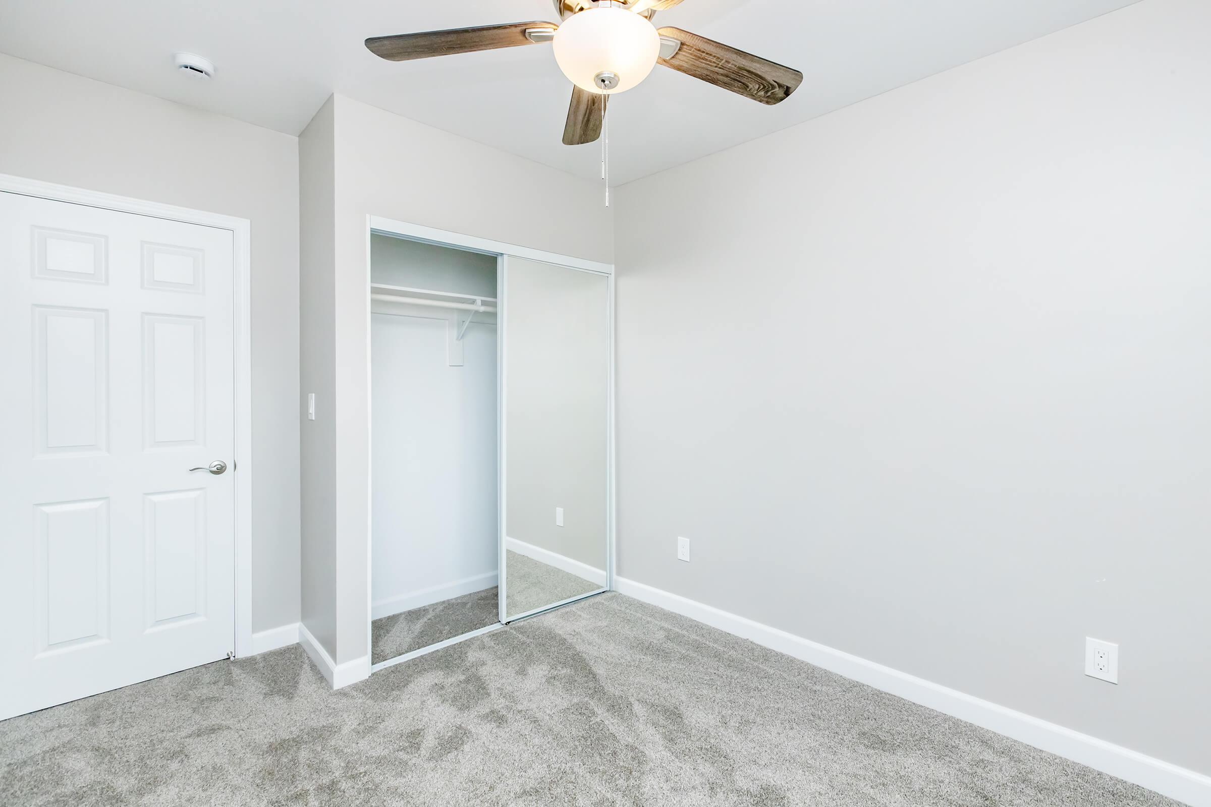 A spacious, neutral-colored bedroom featuring a ceiling fan with wooden blades, a mirrored closet sliding door, and light gray carpeting. The room has a single white door leading to an adjoining space, and the walls are painted in a light shade, creating a bright and airy atmosphere.