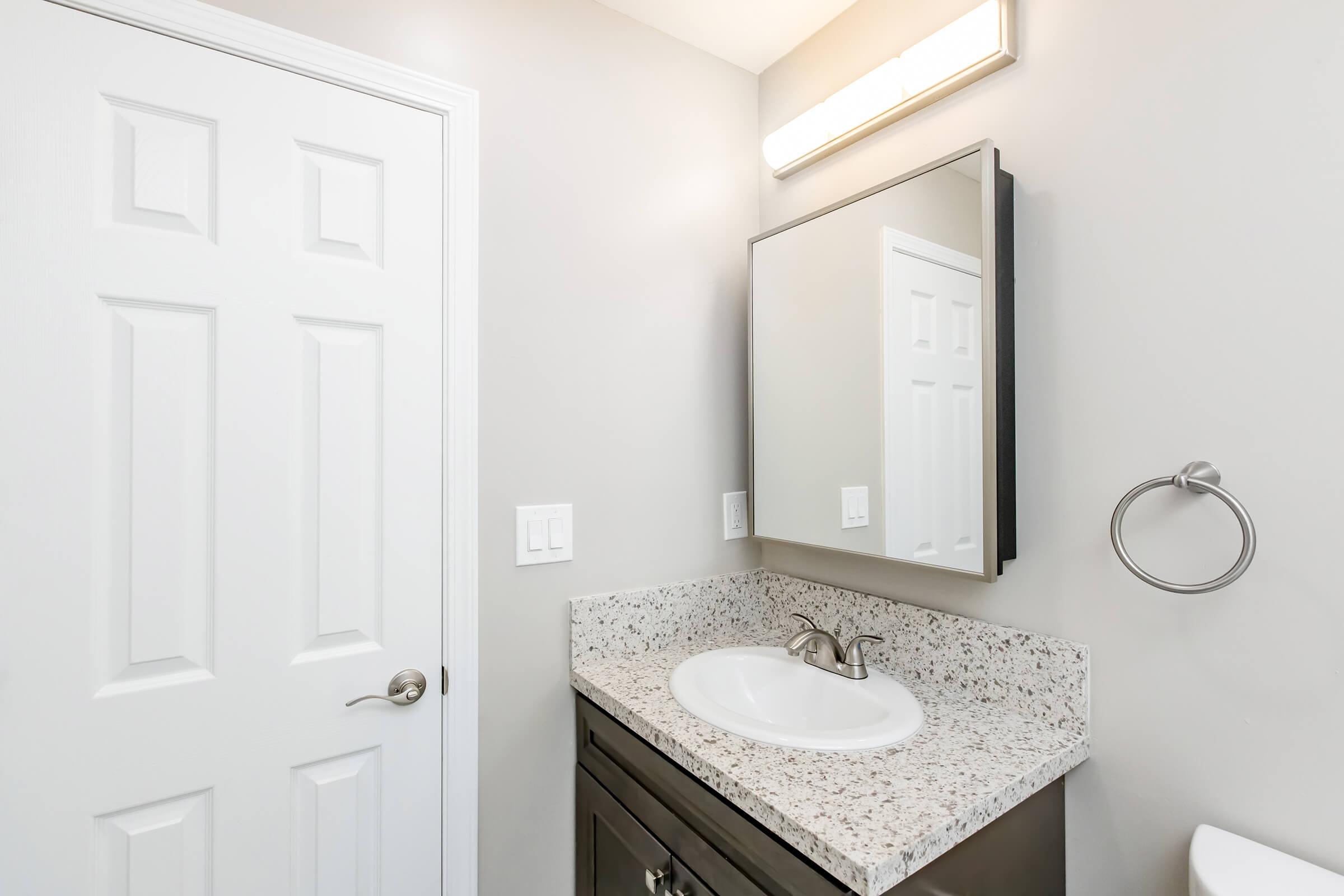 A modern bathroom with a light-colored wall, a mirror above a granite countertop sink, and a towel ring. A white door is closed in the background, and there is a light fixture above the mirror, providing ample lighting for the space.