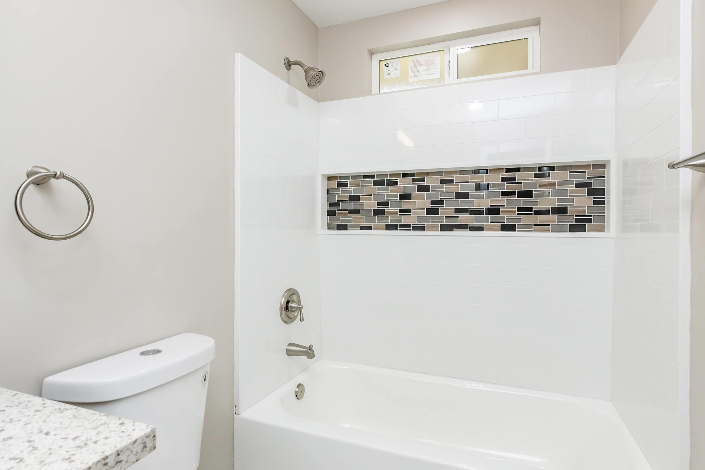 A clean, modern bathroom featuring a bathtub with a tiled accent strip in shades of gray and black. The walls are primarily white, and there's a showerhead mounted above the bathtub. A small window provides natural light, and a towel ring is attached to the wall.