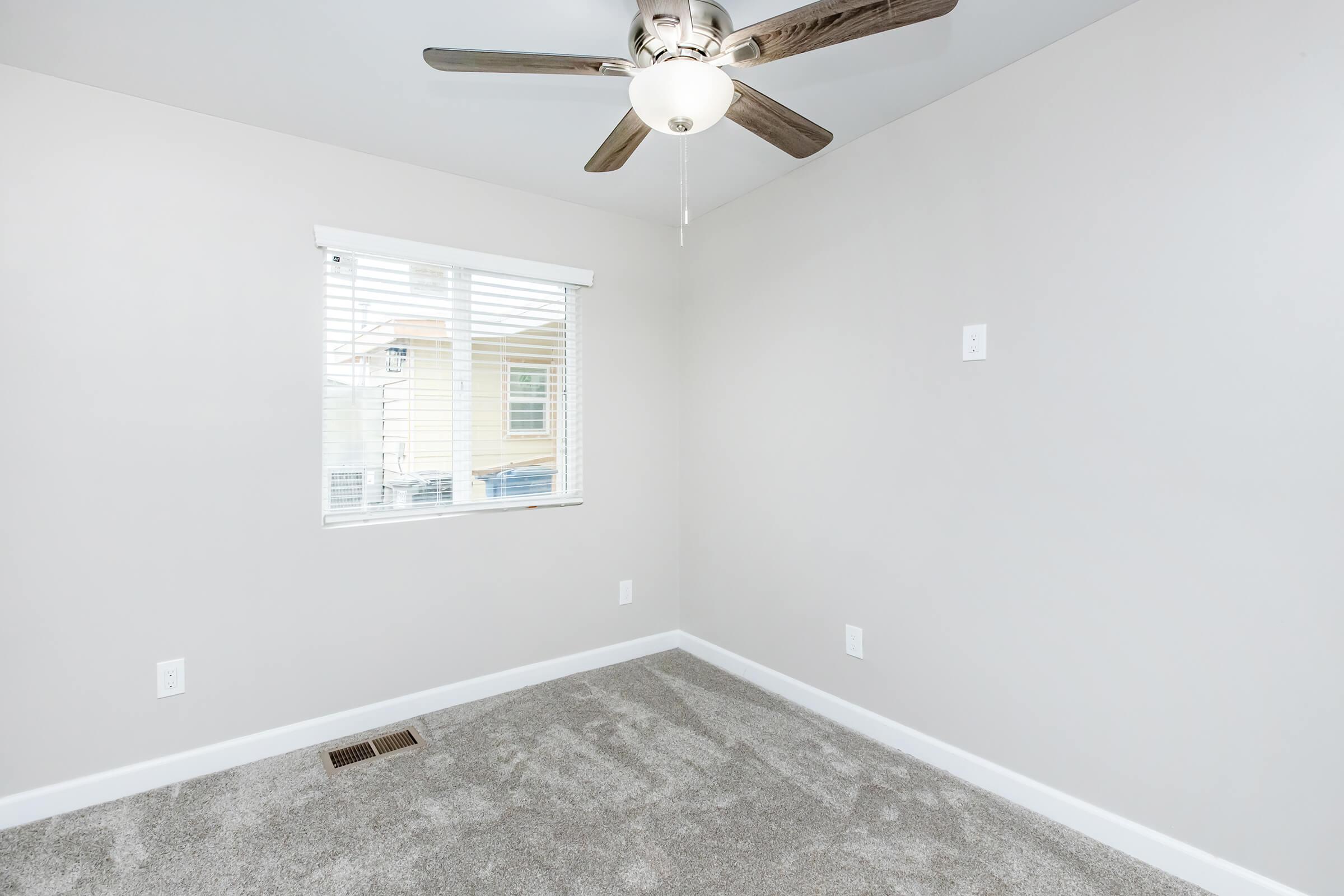 Empty room with light gray carpet and off-white walls. A ceiling fan with a light fixture is mounted on the ceiling. Natural light flows in from a window with white blinds, offering a view outside. There are electrical outlets on the walls, and the floor is clear of furniture.
