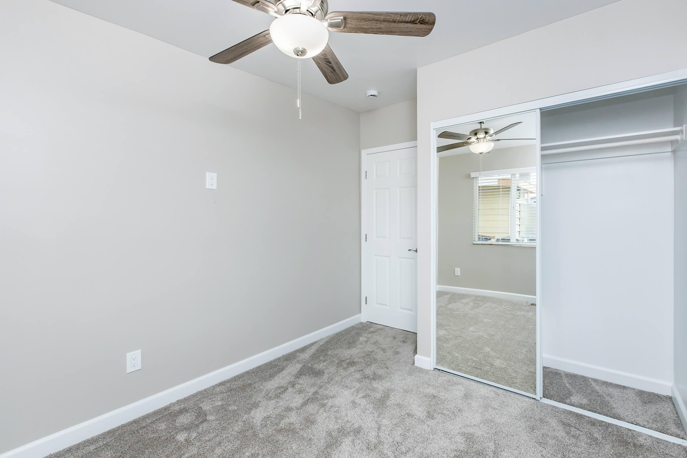 A well-lit, empty room featuring light gray walls, a ceiling fan with wooden blades, and soft gray carpet. A mirrored closet door reflects the room, while a white door leads to another space. The window allows natural light to brighten the area.