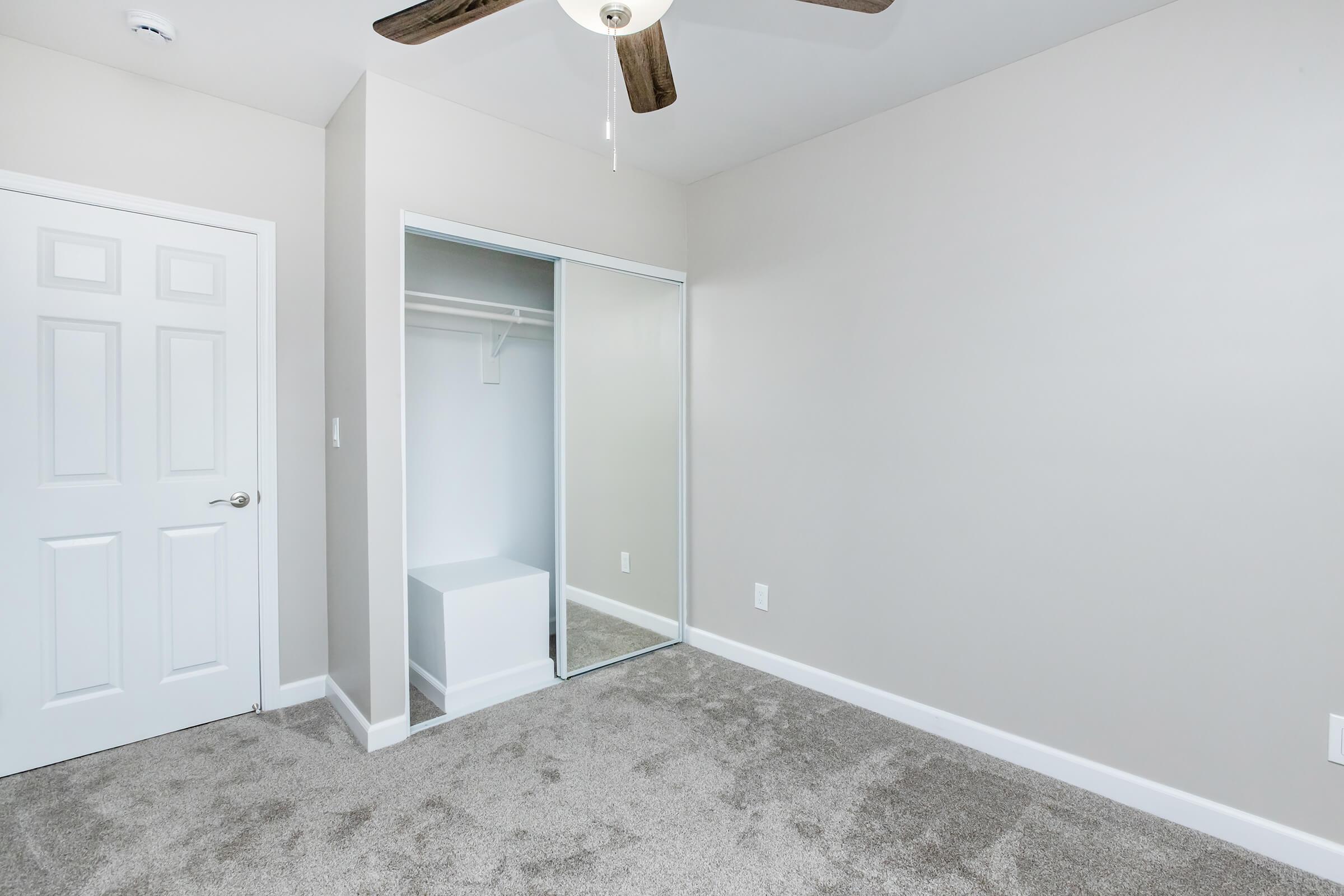 A well-lit, empty room featuring light gray walls and carpet. There is a mirrored sliding closet door on the left, revealing an empty space inside. A ceiling fan is visible above, and a white door leads to another area. The room has a clean and minimalistic aesthetic, suitable for various uses.