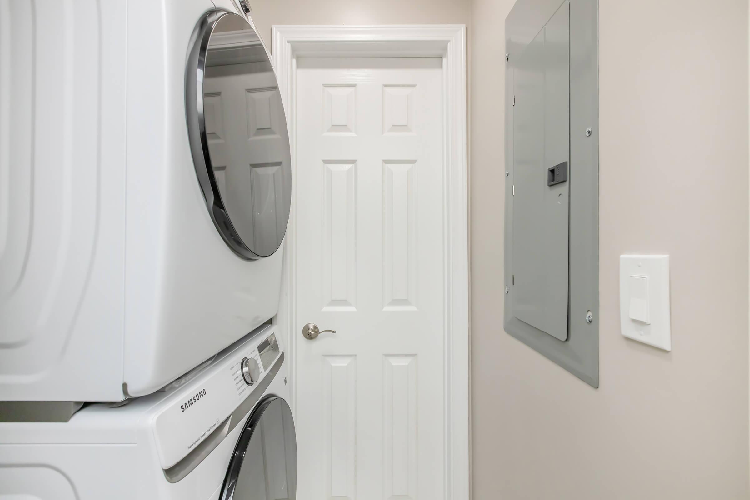 A laundry area featuring a stacked washer and dryer in white, adjacent to a closed white door. To the right, there is a gray electrical panel mounted on the wall, with a light switch positioned nearby. The walls are painted in a neutral tone, creating a clean and organized space.