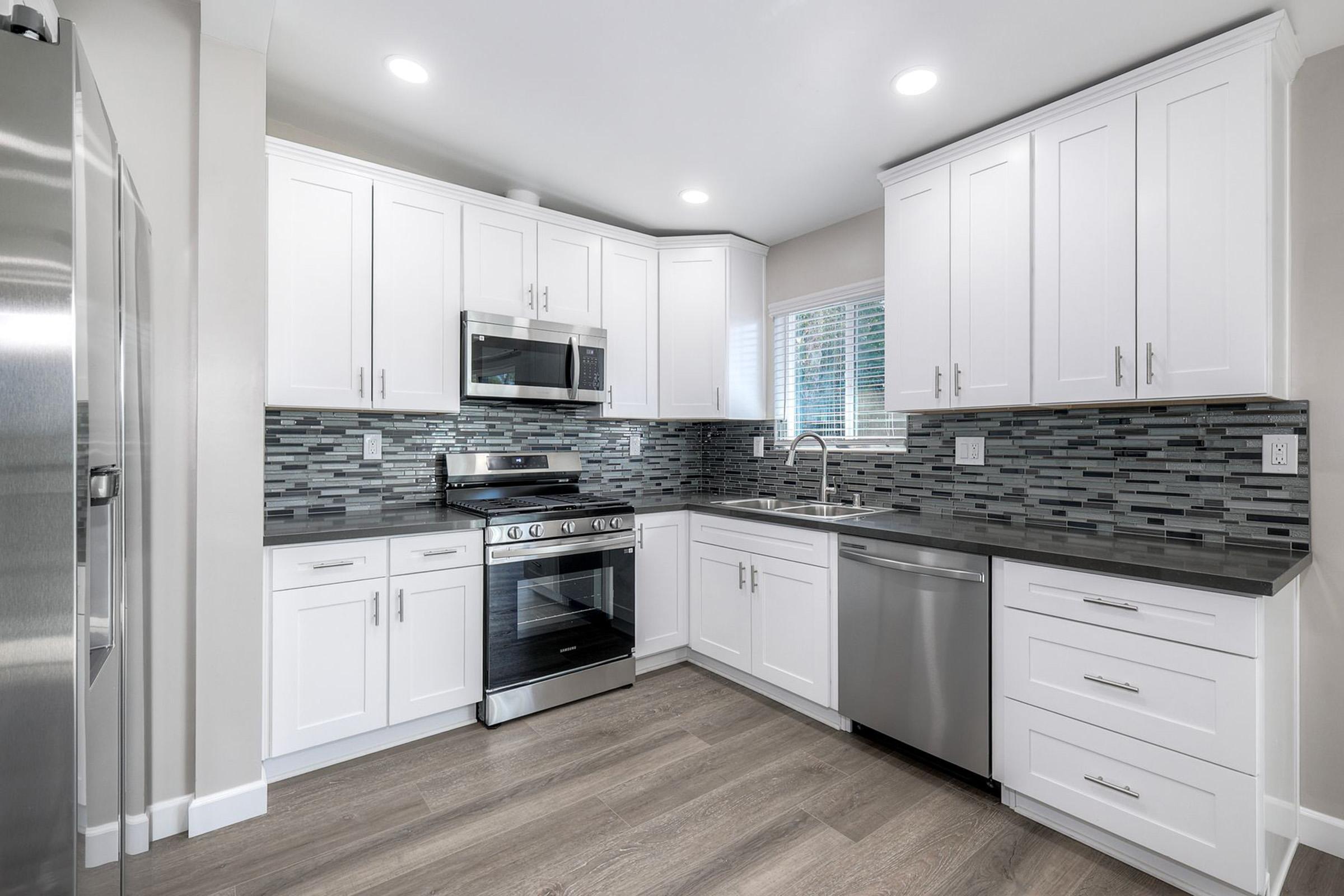 Modern kitchen with white cabinets, stainless steel appliances, and dark countertops. The tile backsplash features gray and black mosaic patterns. Natural light enters through the window, highlighting the wood-like floor. The space appears clean and contemporary, perfect for cooking and entertaining.