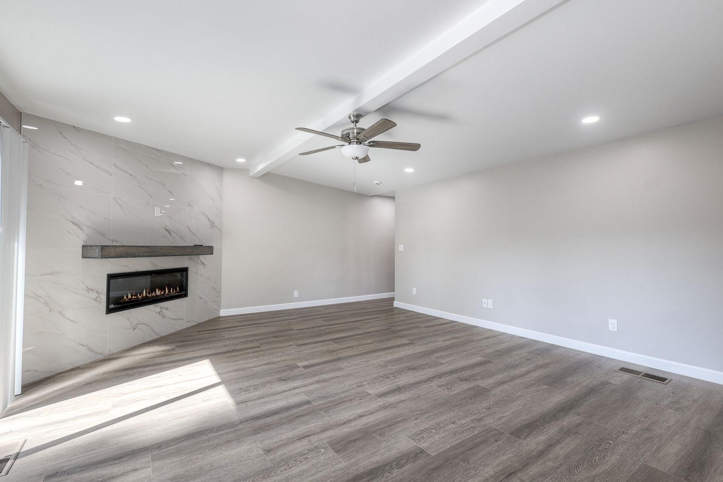 A spacious, modern living room featuring light wood flooring, a ceiling fan, and recessed lighting. The wall contains a sleek, modern fireplace with a marble surround, and large windows allow natural light to enter, highlighting the neutral-colored walls.