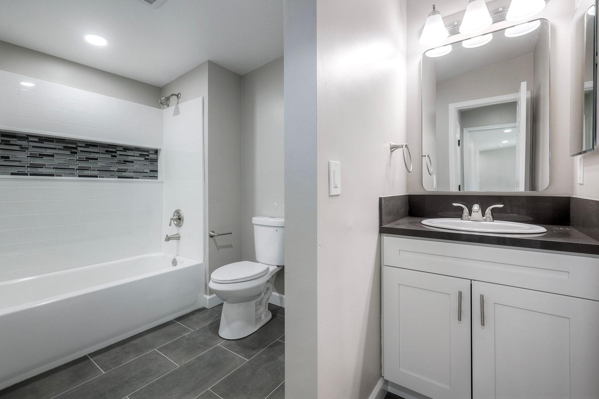 Modern bathroom featuring a white bathtub with a tiled accent wall, a toilet, and a sleek vanity with a dark countertop. Bright lighting from overhead fixtures enhances the space, which has neutral-colored walls and gray floor tiles, creating a clean and inviting atmosphere.
