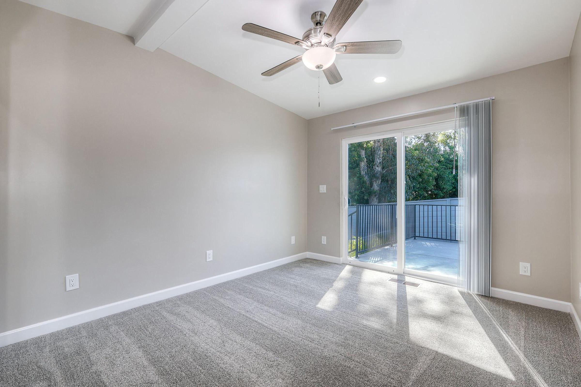 A bright and spacious room featuring a ceiling fan, large window with sliding glass doors, and plush gray carpeting. The walls are painted a soft neutral color, and there is a balcony visible through the sliding doors, offering a view of outdoor greenery.