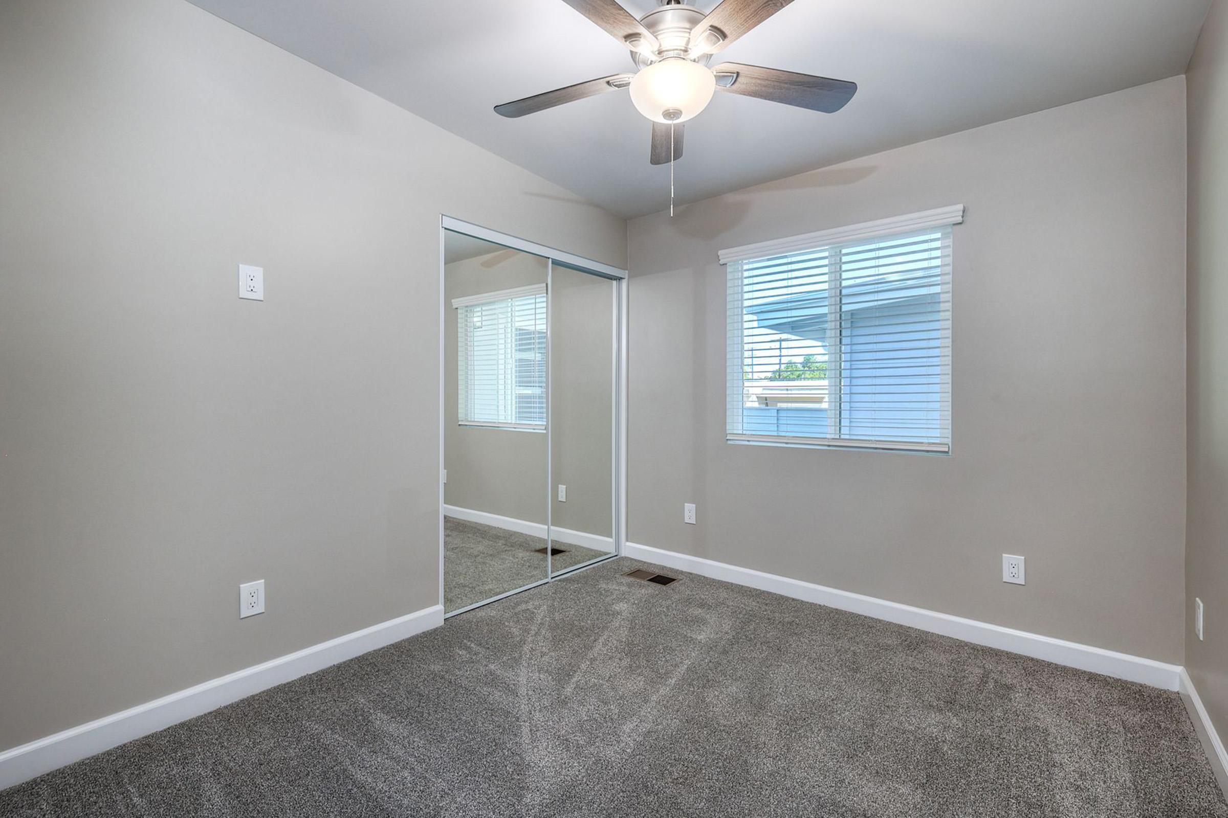 A well-lit, empty bedroom featuring light gray walls, a ceiling fan, and a large mirrored closet. The floor is covered in soft gray carpet, and there's a window with blinds allowing natural light to enter.