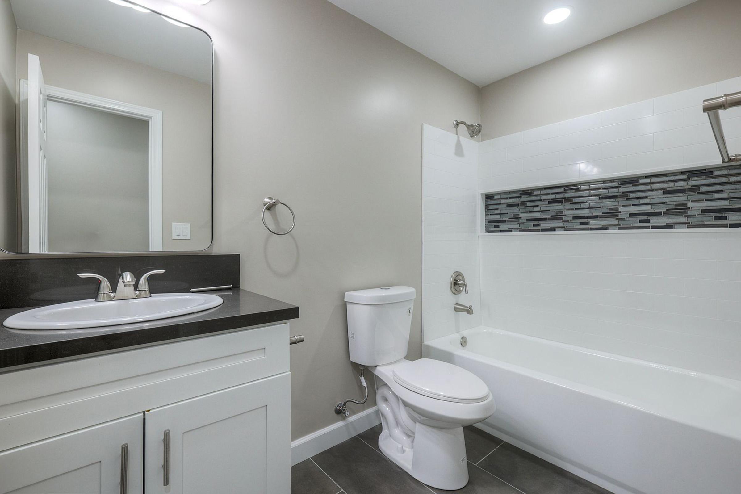 A modern bathroom featuring a white bathtub with a shower, a toilet, and a sink with a dark countertop. The walls have a light neutral color, and there's a decorative tile strip in the bathtub area. A large mirror and towel holder are also visible, with bright overhead lighting illuminating the space.