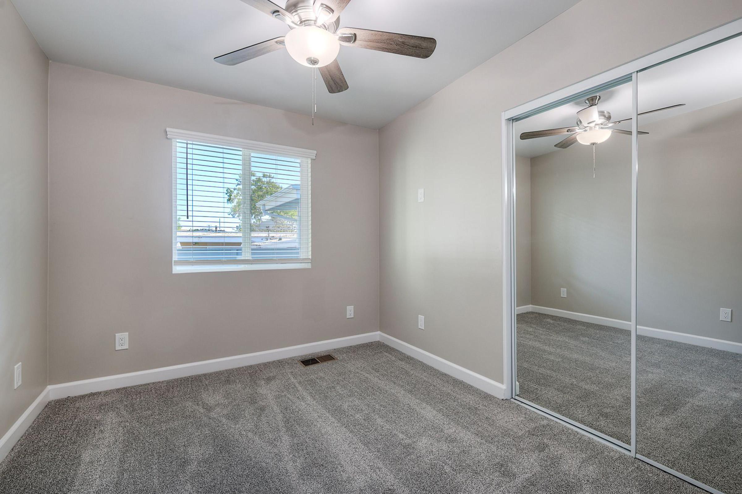 A well-lit, empty room featuring light gray walls and new carpet. It has a window with blinds on the left, allowing natural light in. There are two ceiling fans, one in the center and one near the mirrored closet doors, which reflect the room's interior. The overall ambiance is clean and modern.