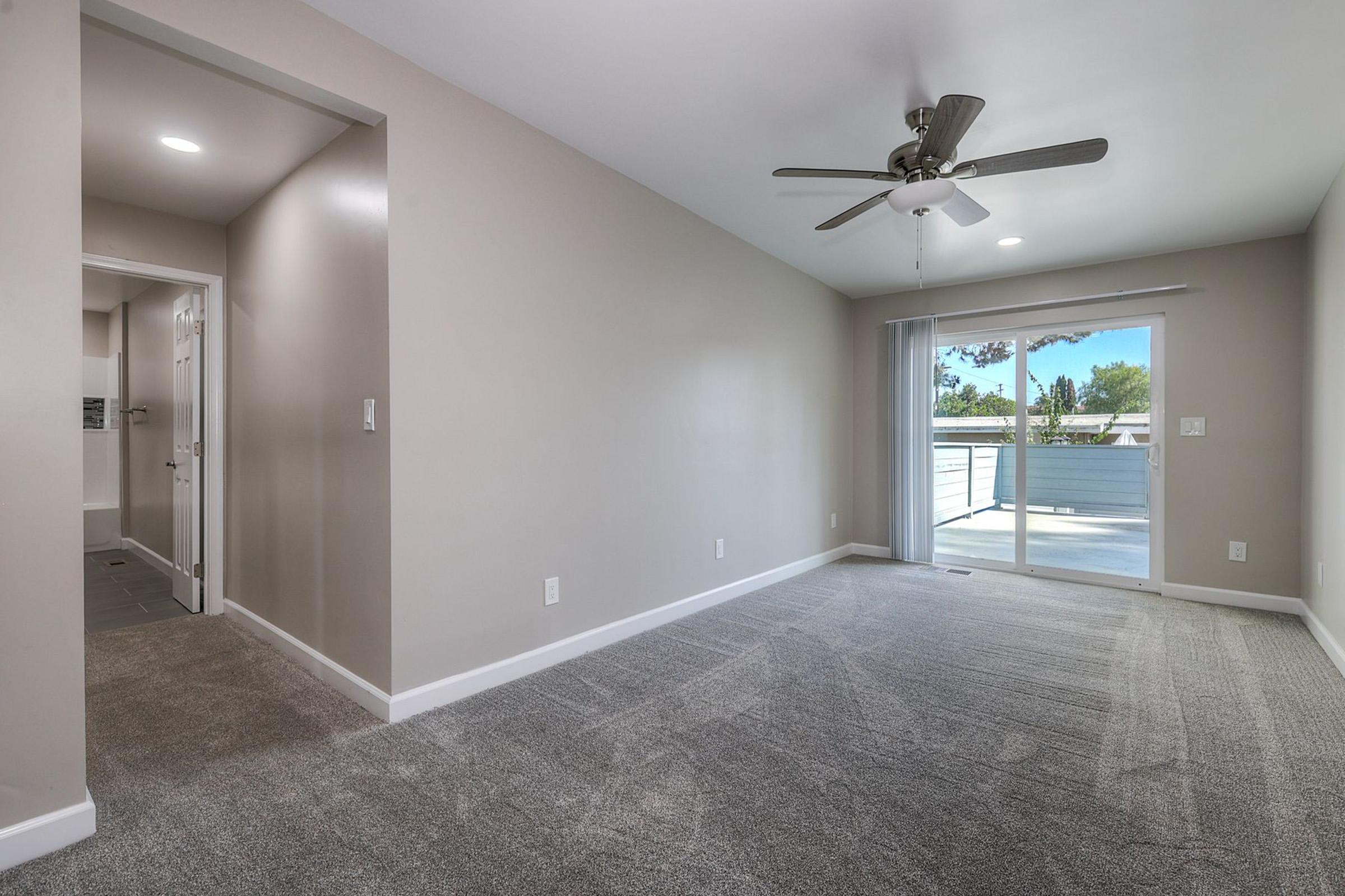 A spacious, light-filled living room featuring light gray walls and plush carpeting. A ceiling fan hangs from the ceiling, and large sliding glass doors lead to a balcony or outdoor area. To the left, a doorway opens into a hallway, with bright lighting enhancing the open atmosphere.
