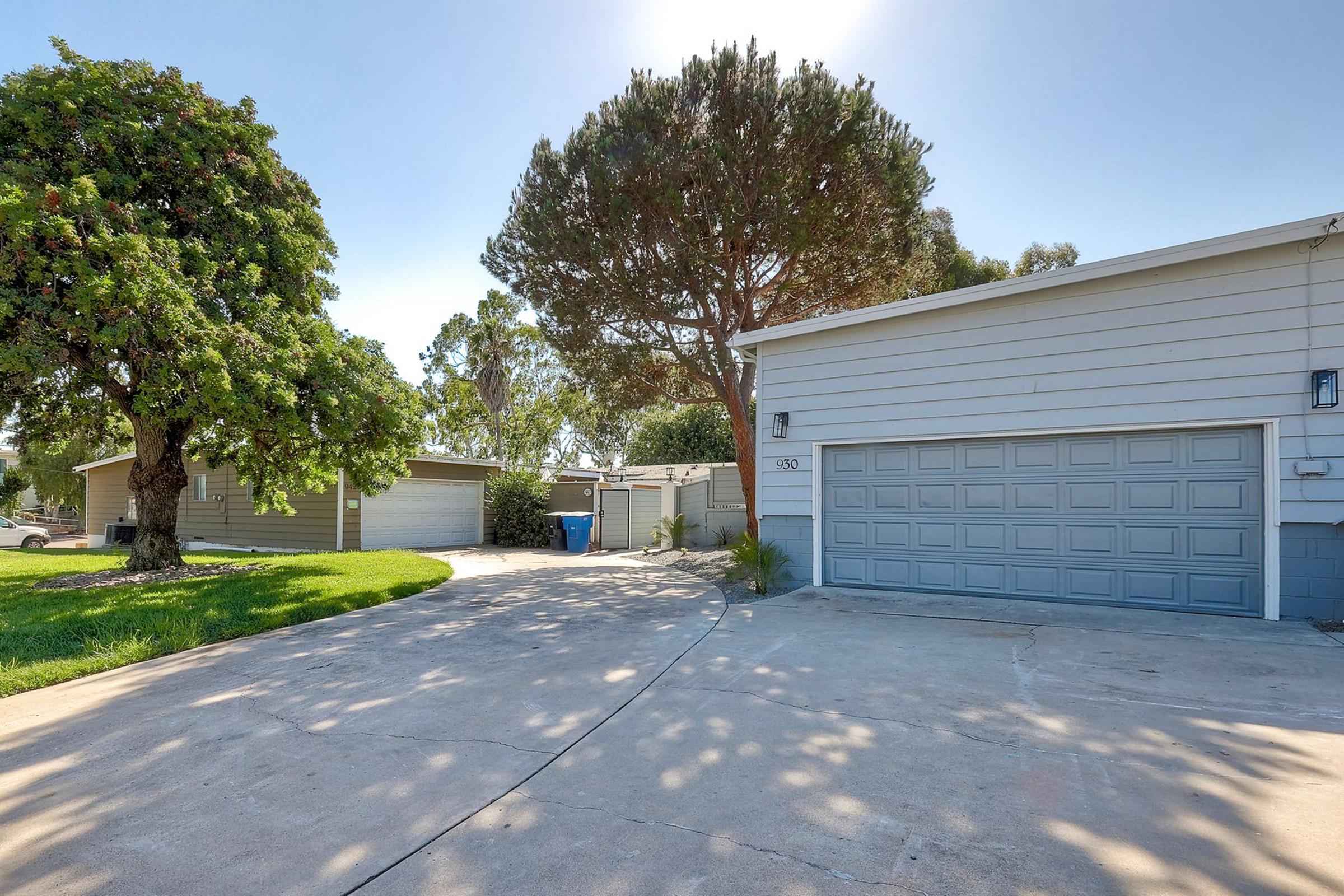 A residential property featuring a driveway leading to a garage, surrounded by well-maintained lawns and trees. The scene includes a blue trash bin near the garage and a clear sky illuminating the area.