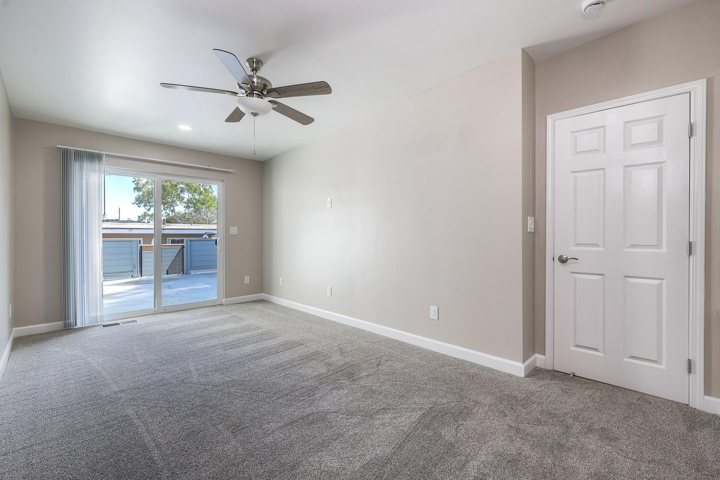 Spacious and bright room featuring light gray walls, a ceiling fan, and a large sliding door that opens to a balcony. The floor is covered with soft, light-colored carpet, and there is a closed white door on the right. The overall ambiance is modern and inviting, suitable for various uses.