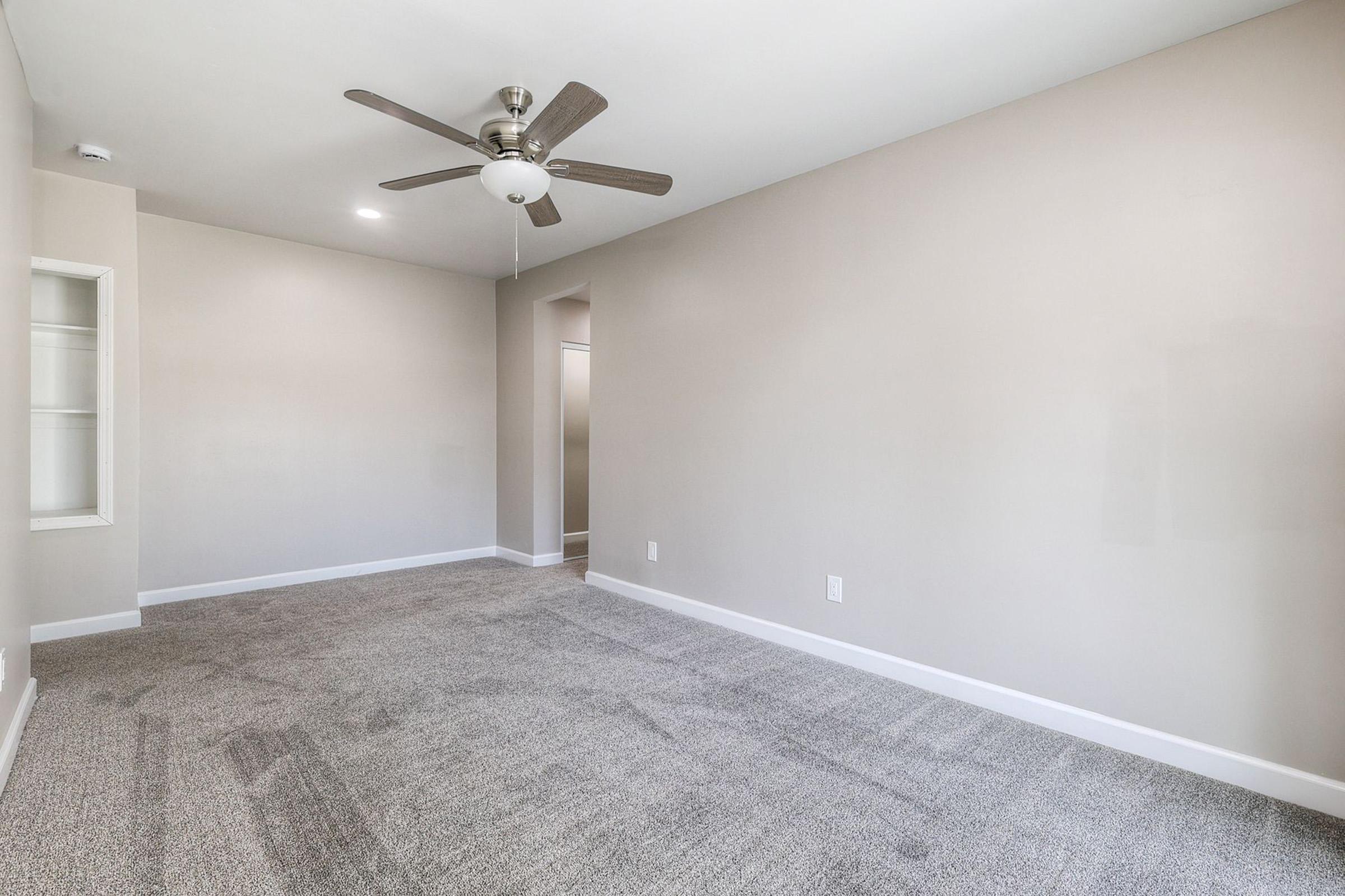 A spacious, empty room with light gray carpet flooring, a ceiling fan with light fixture, and neutral-colored walls. There is a small nook with built-in shelves on the left and an open doorway leading to another room in the background.