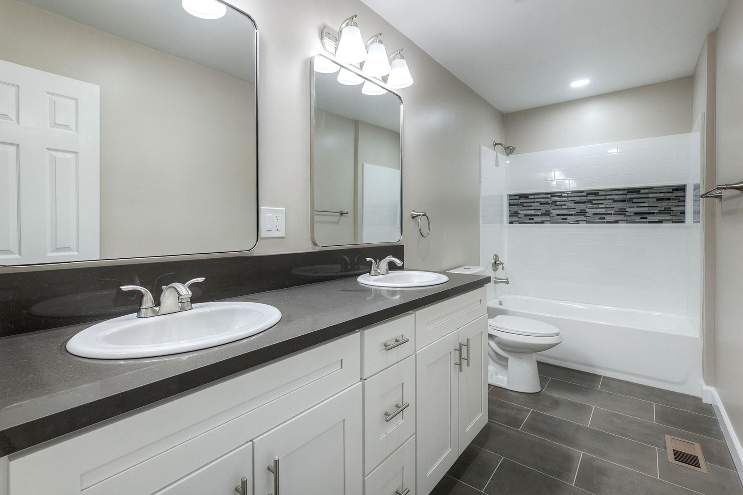 Modern bathroom featuring a double vanity with two sinks and large mirrors above, along with a bathtub surrounded by white tiles and a decorative gray mosaic accent. The space has neutral wall colors, sleek black countertop, and contemporary lighting fixtures, creating a bright and clean atmosphere.