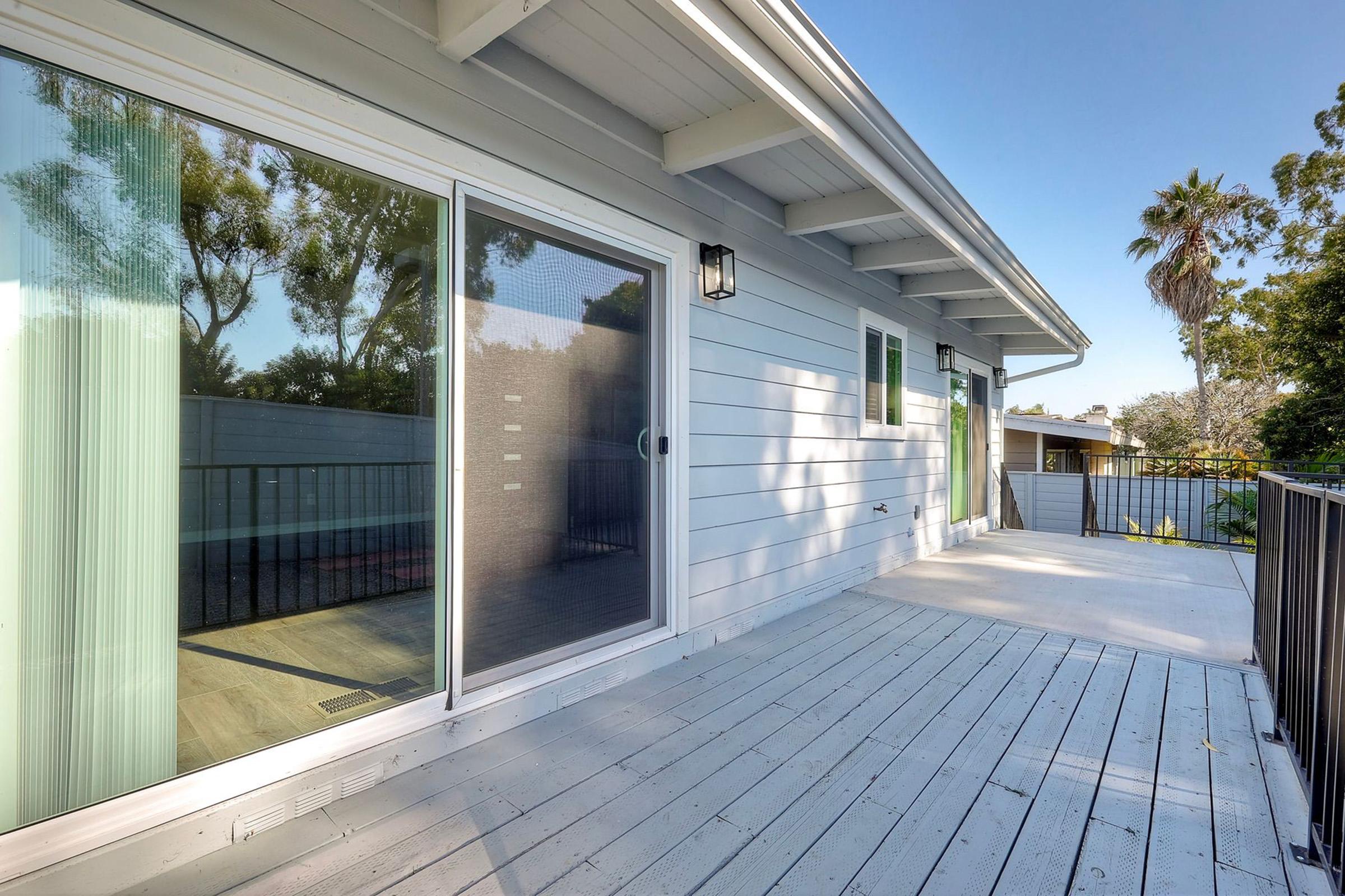 A modern deck area with light-colored wooden flooring and sliding glass doors, showcasing a spacious outdoor space. Lush green trees and a palm are visible in the background, indicating a sunny outdoor environment. The house has a contemporary design with clean lines and ample natural light.