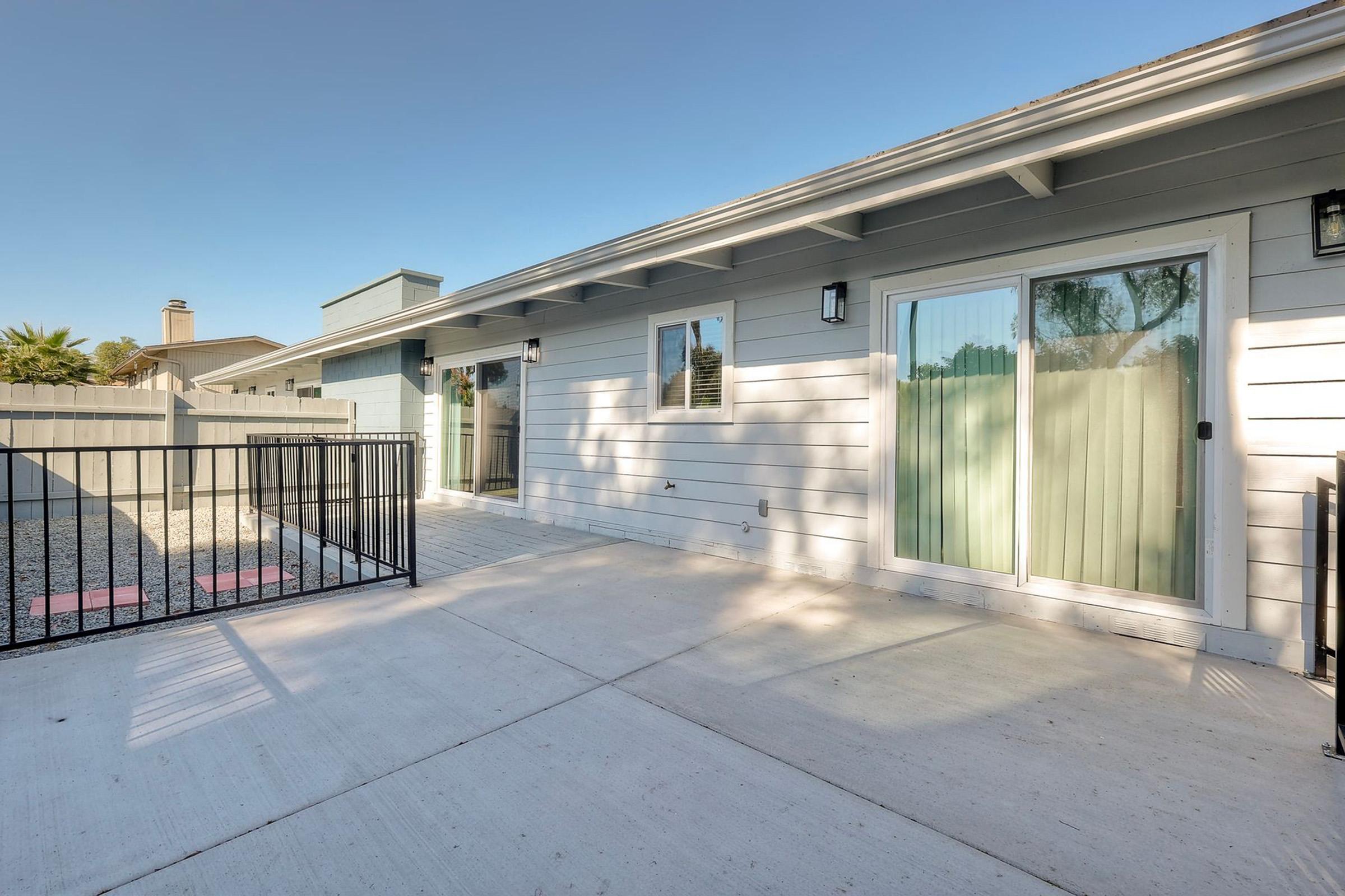 Exterior view of a modern single-story house with a light-colored siding and large sliding glass doors. The patio area is paved, with a fenced yard to the side and clear blue sky overhead. The surrounding area includes a neighboring house and green landscaping in the background.