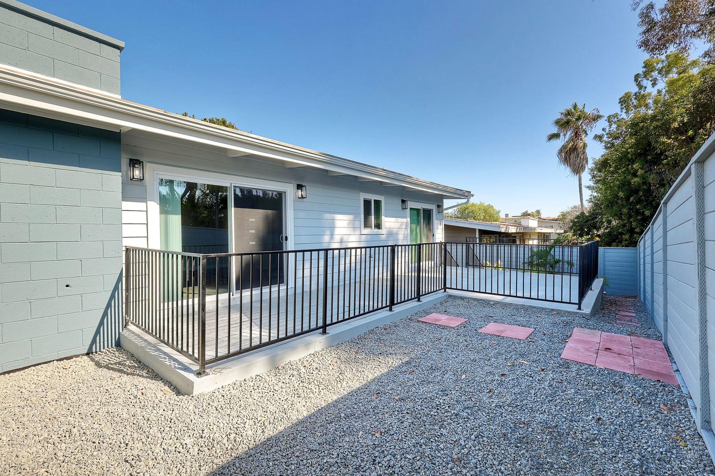 A modern outdoor space featuring a small patio area with a gravel surface, bordered by a black metal railing. There are two sliding doors leading to the interior, and a few concrete stepping stones are placed on the ground. Lush greenery and a palm tree are visible in the background, reflecting a sunny day.