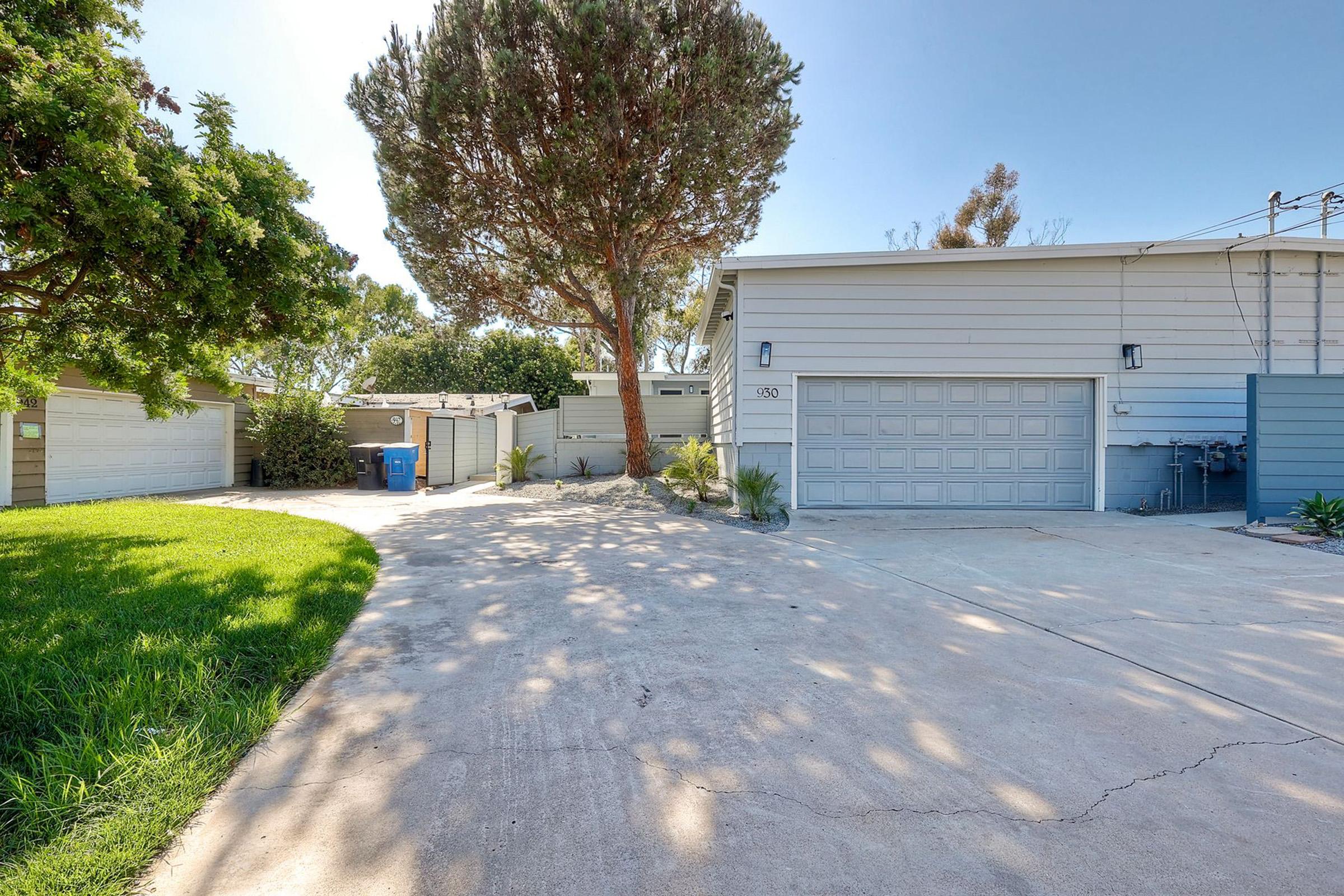 A residential property featuring a gray house with a garage, situated on a driveway. Lush green grass and a tree are visible, alongside a blue trash bin and a fenced yard. The scene appears sunny with clear skies and minimal landscaping details.