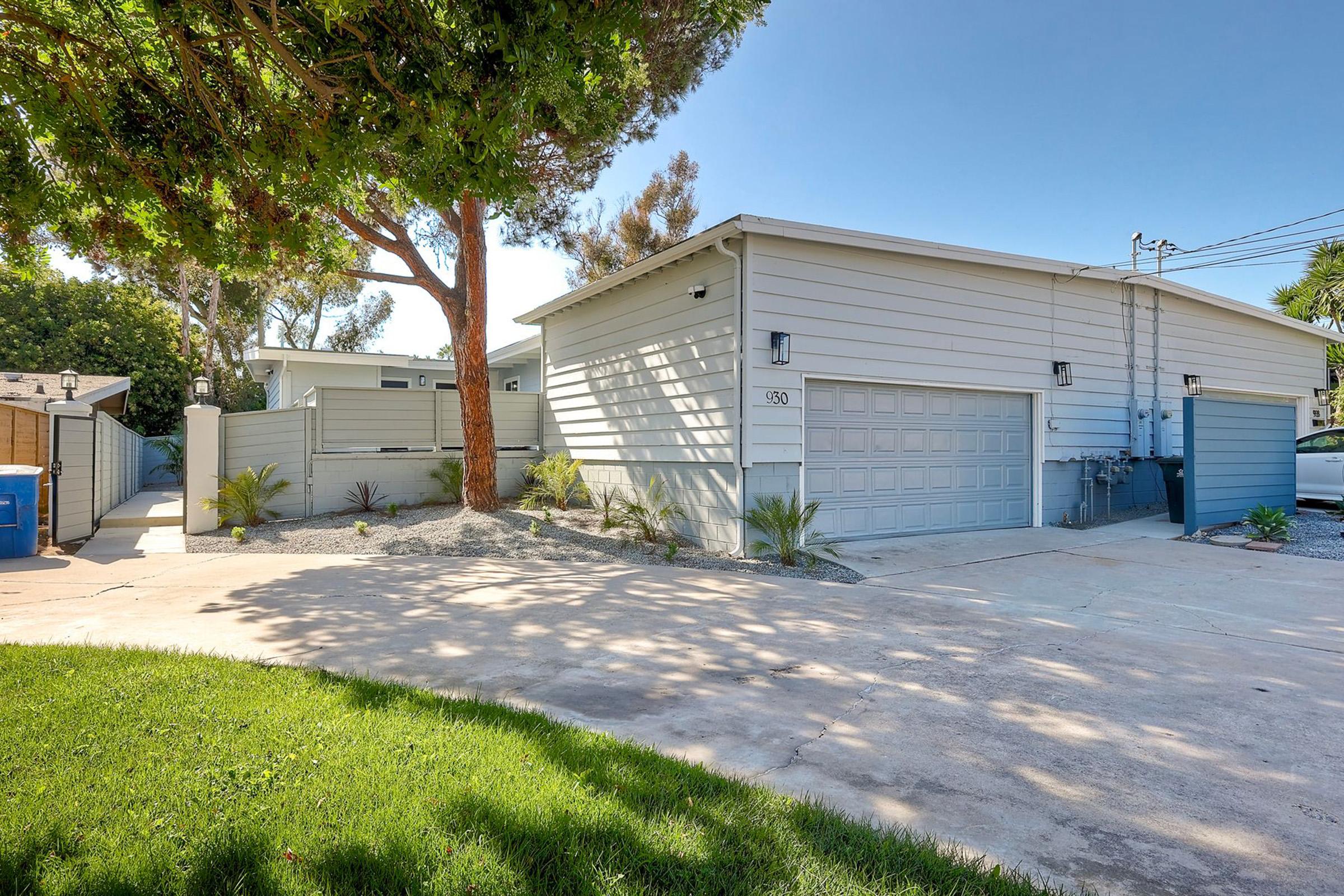 A modern, single-story house with a light gray exterior, featuring a garage and landscaped yard with green grass and palm plants. In the background, there is a tall tree and a blue trash can, with utility lines visible against a clear sky. The driveway is paved and leads to the garage.