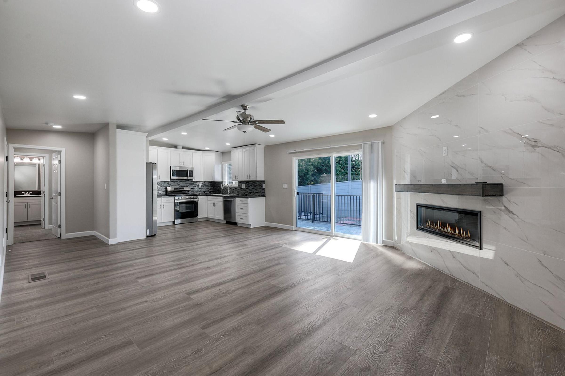 Spacious modern living room with hardwood flooring, large windows, and a sliding glass door leading to an outdoor area. The room features a sleek white fireplace and a stylish kitchen with dark countertops and stainless steel appliances. Bright, neutral-colored walls enhance the open layout.