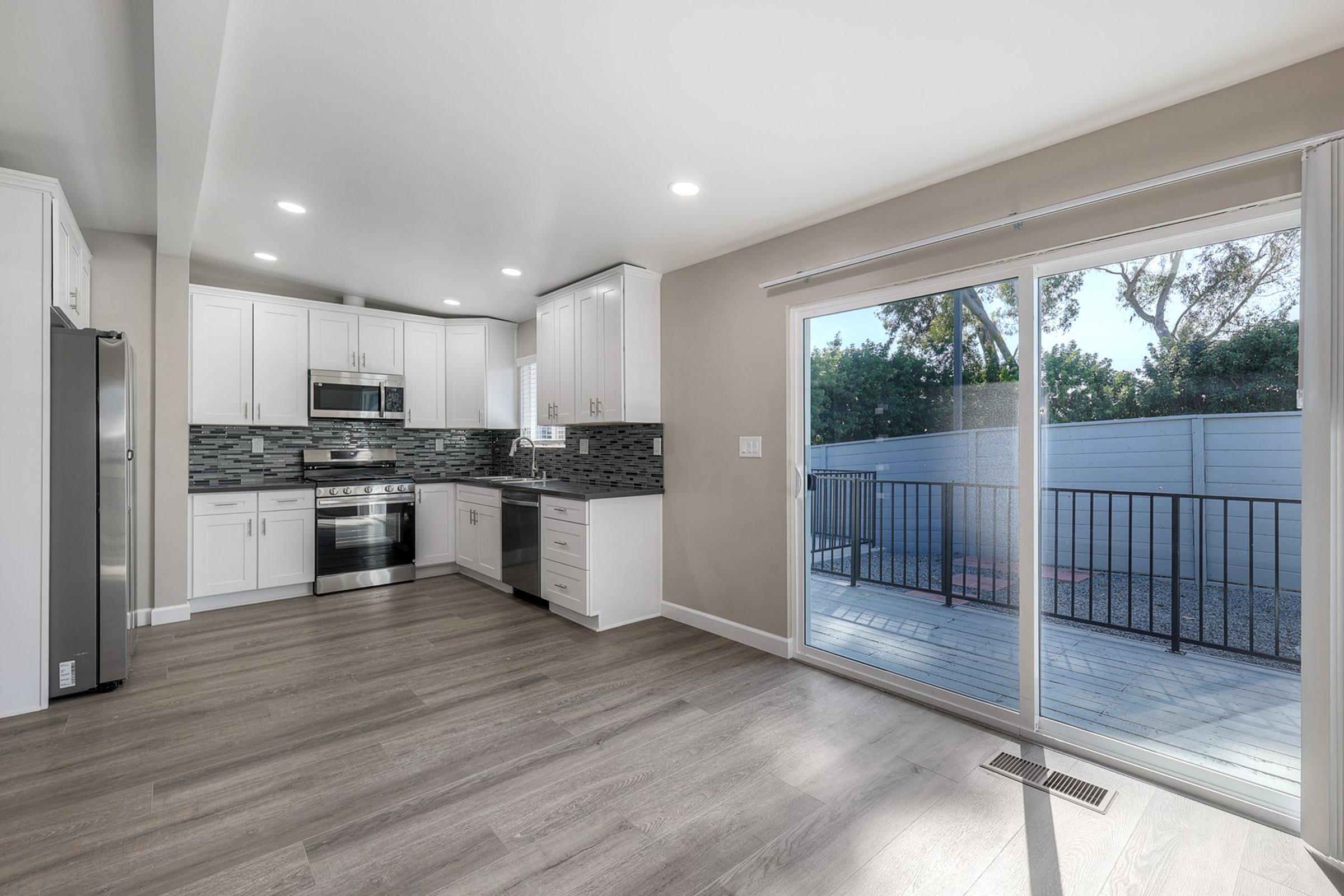 A modern kitchen featuring white cabinetry, stainless steel appliances, and a tiled backsplash. The space includes a large window with a sliding door leading to a patio area. The floor is made of light wood, creating a bright and airy atmosphere with ample natural light.
