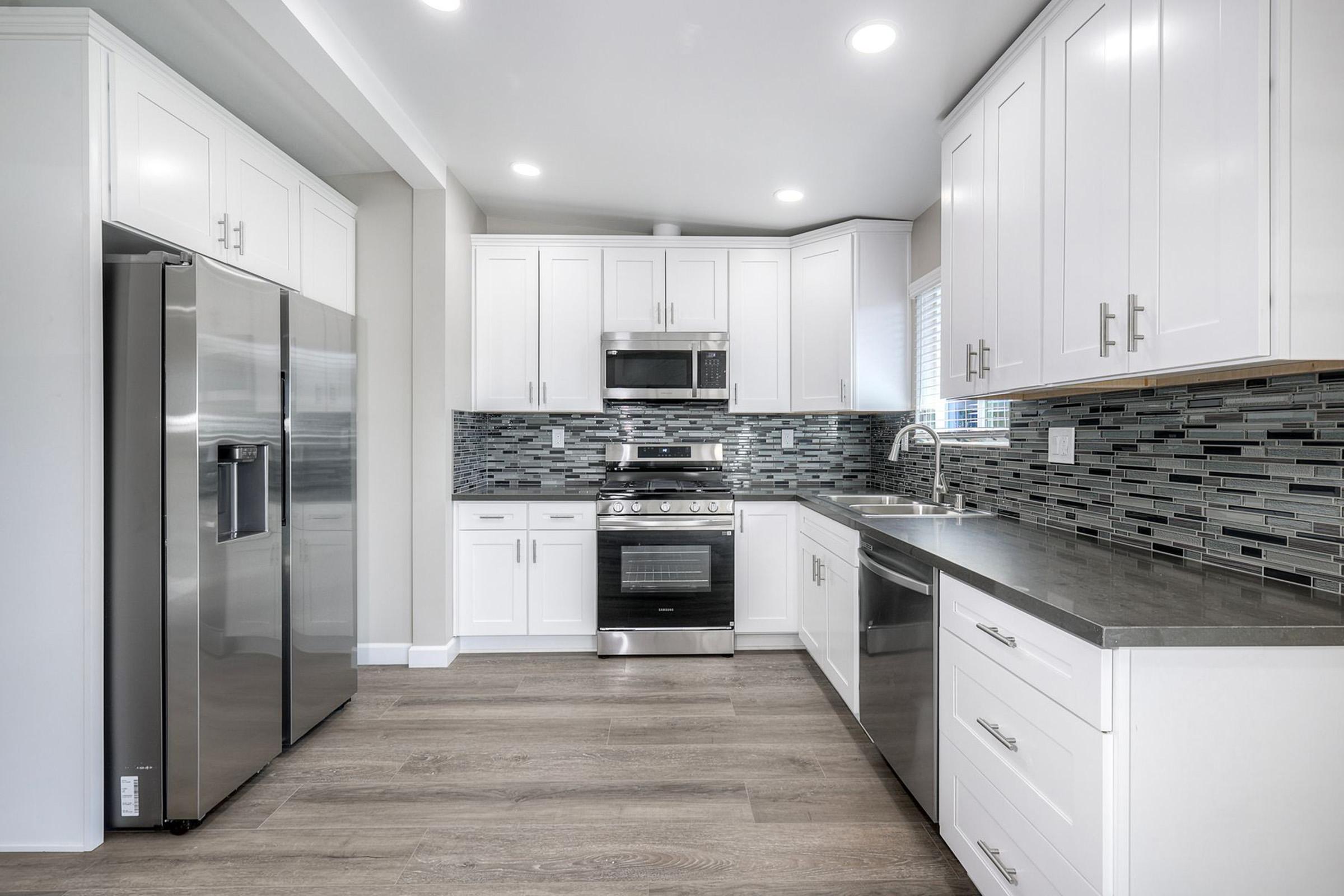 Modern kitchen featuring stainless steel appliances, including a refrigerator and stove. The cabinetry is white with chrome handles, and a stylish backsplash of blue and gray tiles complements the space. Natural light streams in, highlighting the sleek countertops and wood-like flooring.