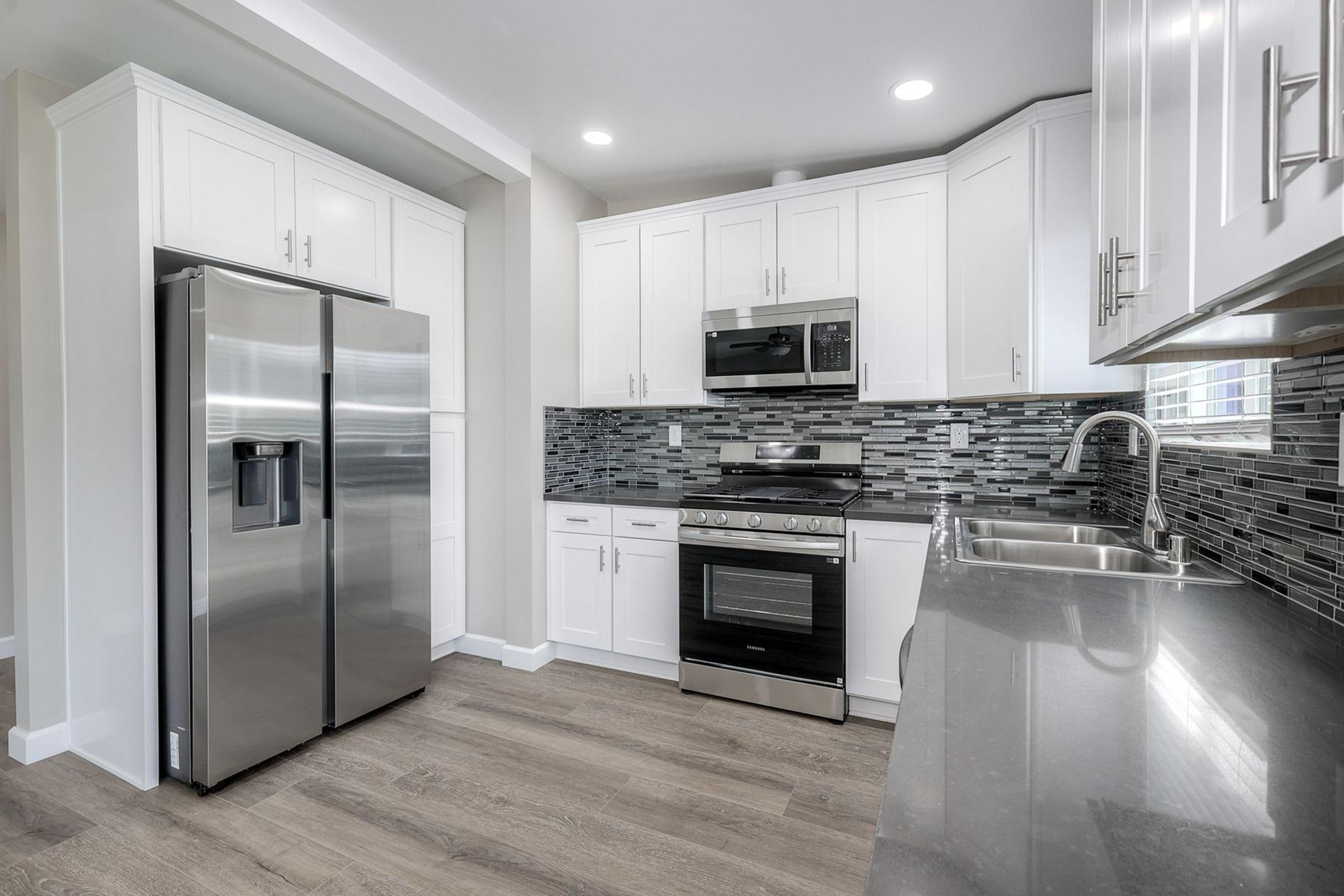 Modern kitchen featuring stainless steel appliances, including a refrigerator and oven, with white cabinetry and a sleek gray countertop. The backsplash consists of a mosaic of gray tiles, and the flooring is light wood. Bright lighting enhances the contemporary design.
