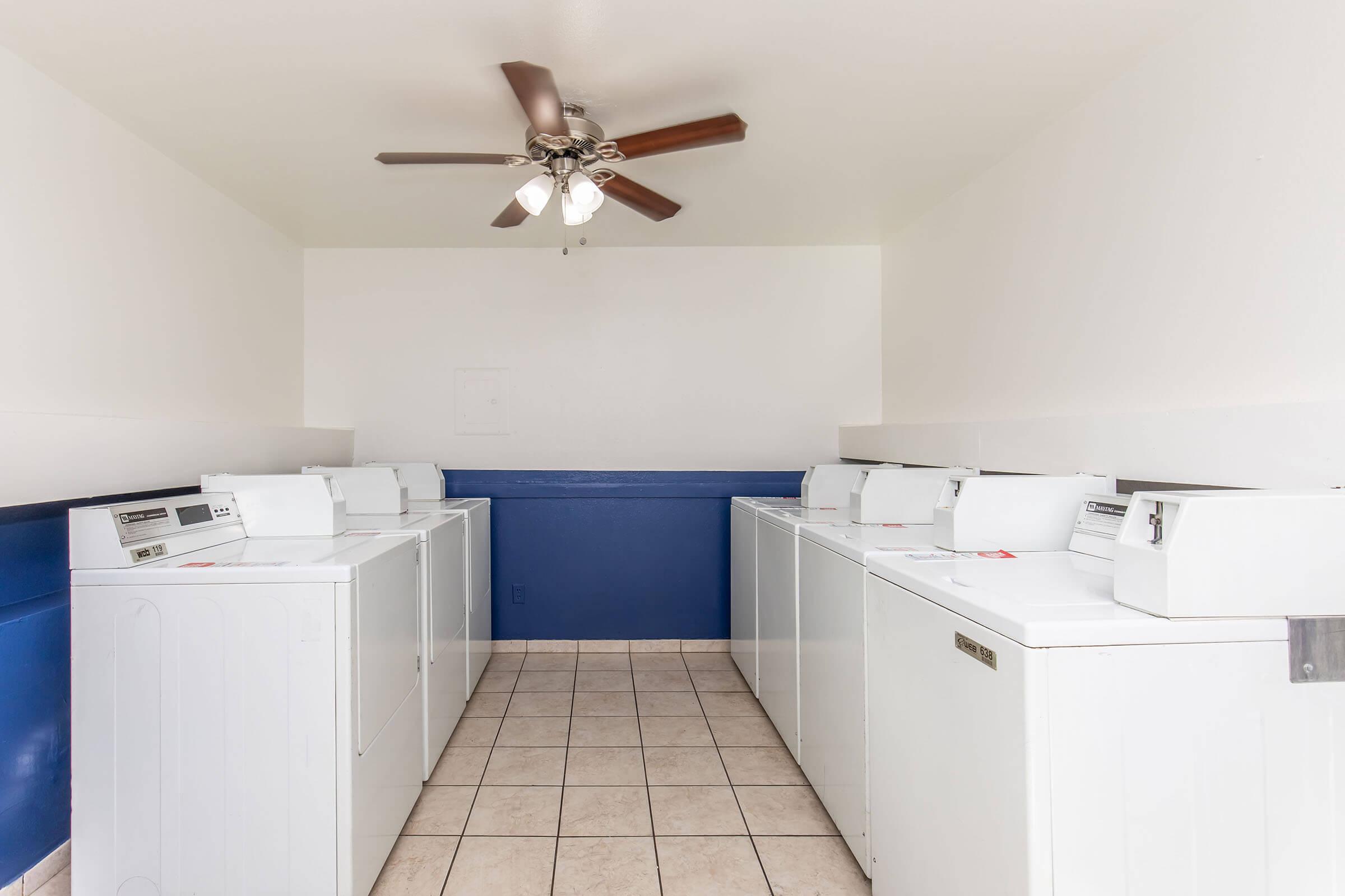 A clean and well-lit laundry room featuring several white washing machines and dryers arranged in rows. The room has a tiled floor, a ceiling fan, and blue painted walls, creating a neat and functional space for laundry activities.
