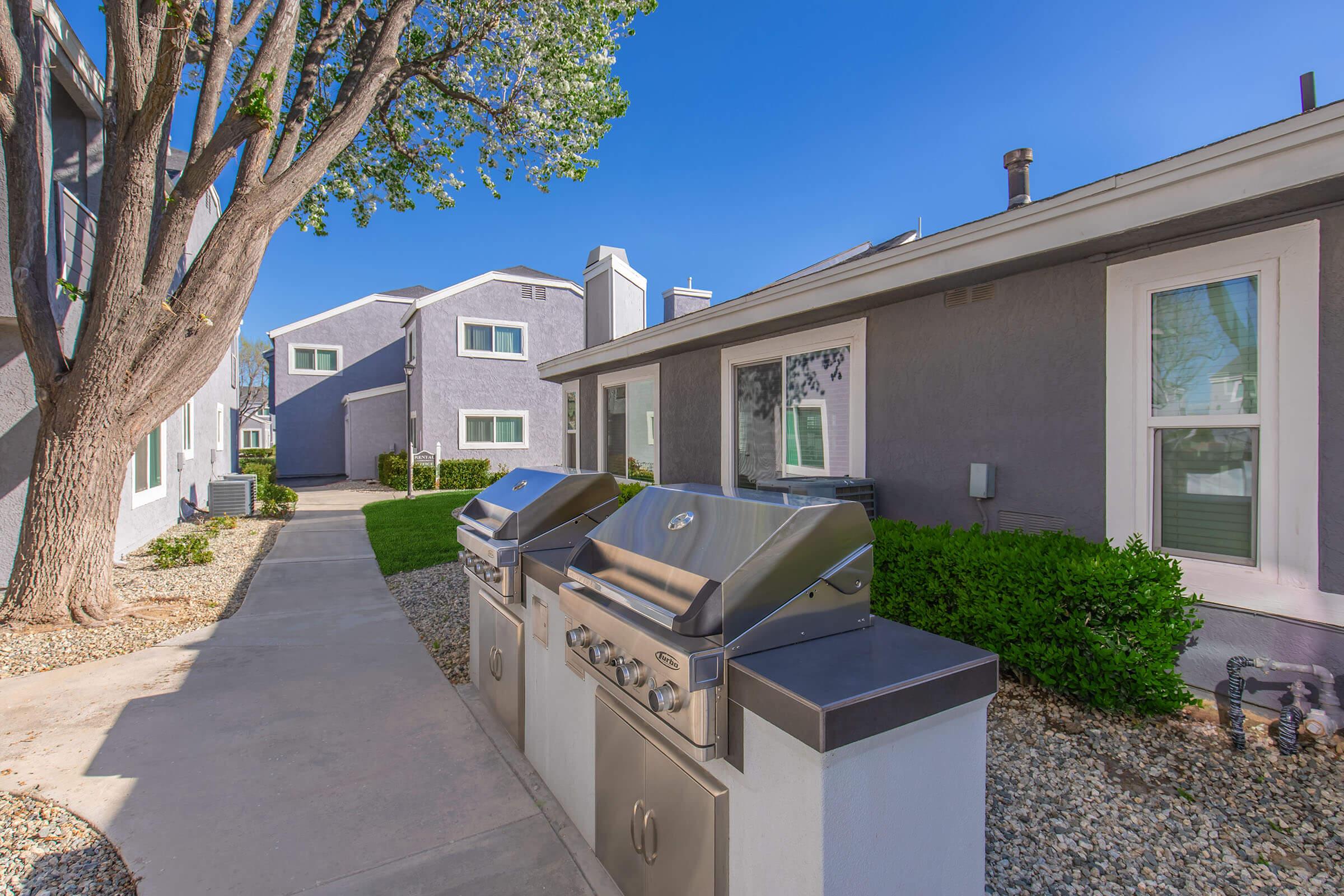 Outdoor area featuring stainless steel grills placed along a pathway, surrounded by green lawns and shrubs. Grey apartment buildings are visible in the background under a clear blue sky. The scene suggests a well-maintained residential environment ideal for outdoor cooking and gatherings.