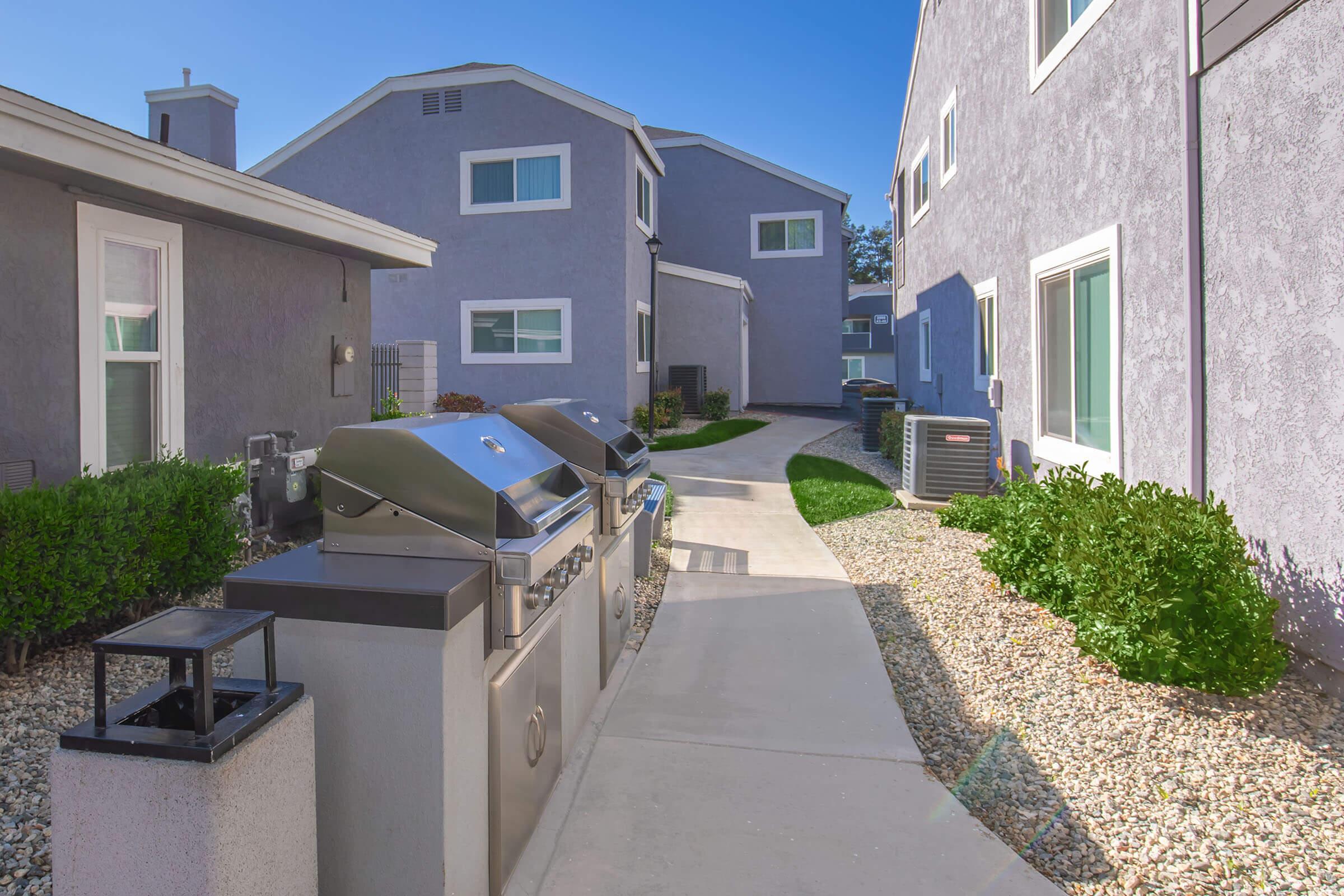 Pathway lined with modern two-story buildings, featuring barbecue grills and landscaped greenery. The bright blue sky adds natural light to the scene, creating an inviting outdoor space.
