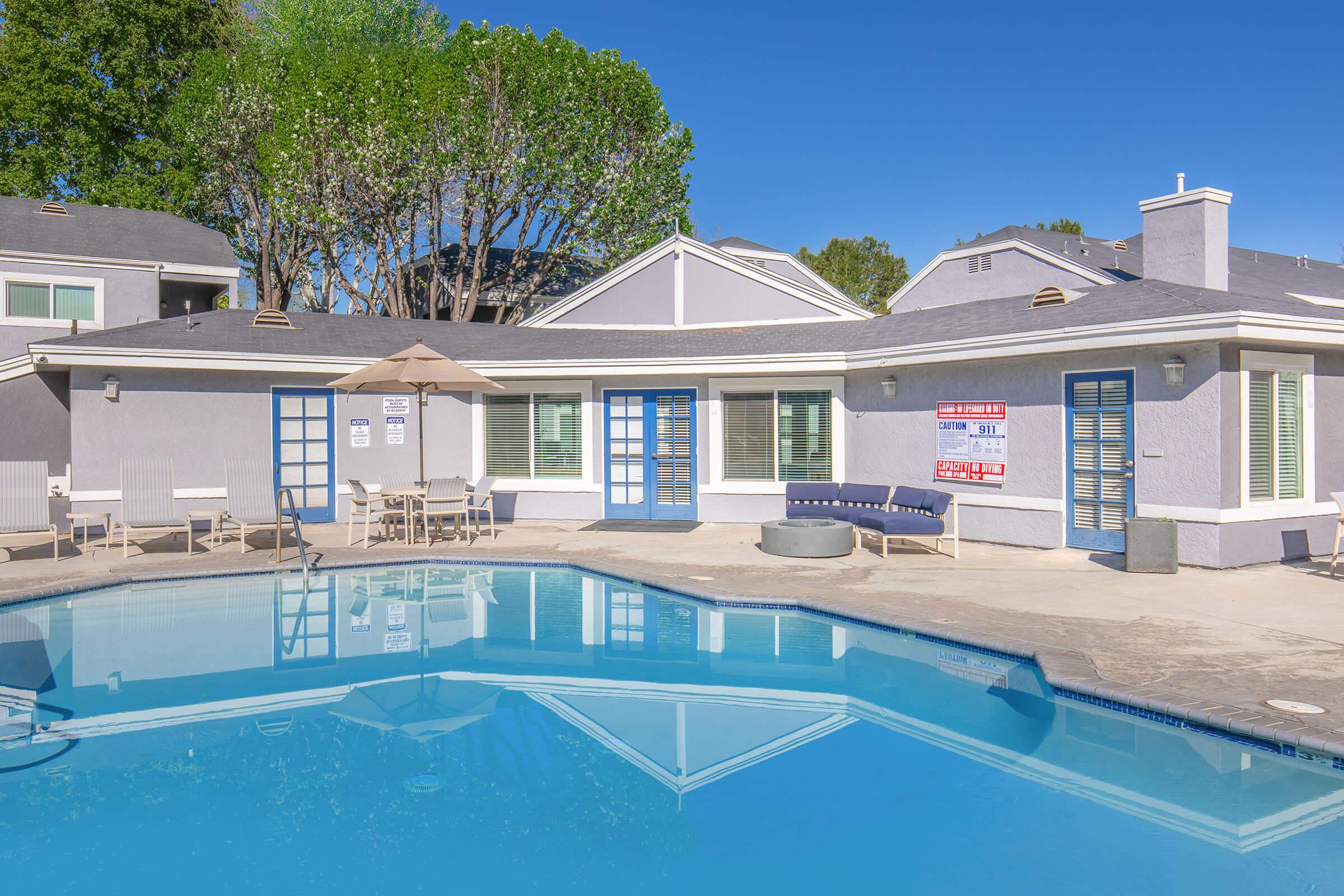 A bright swimming pool surrounded by a patio area with lounge chairs and an umbrella. In the background, there is a well-maintained building with blue doors and windows, framed by lush green trees under a clear blue sky.